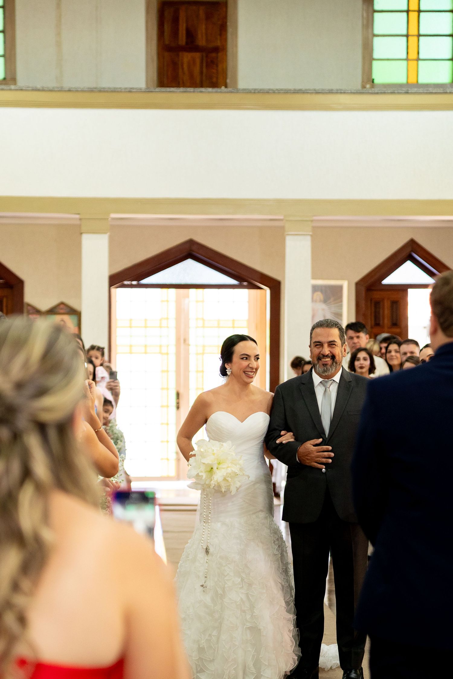 Bride walks down church aisle with father, smiling, holding white bouquet, guests watching during wedding ceremony.
