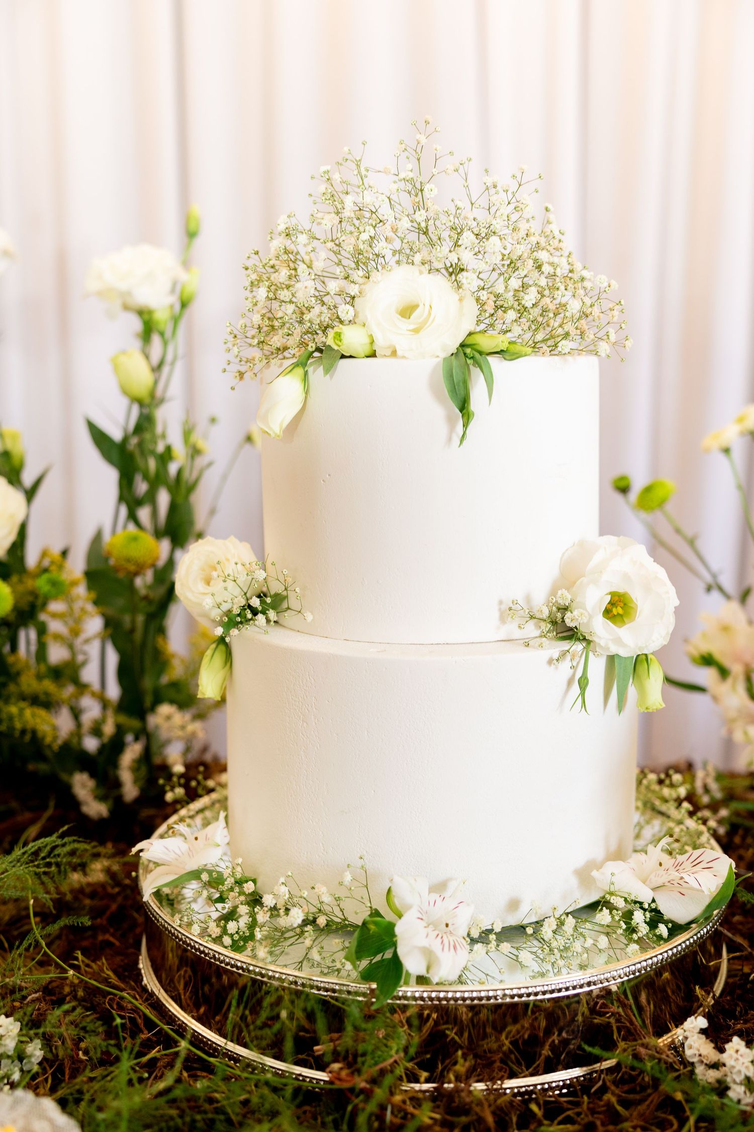 Elegant two-tier white wedding cake adorned with fresh white roses and baby's breath flowers on a silver stand.