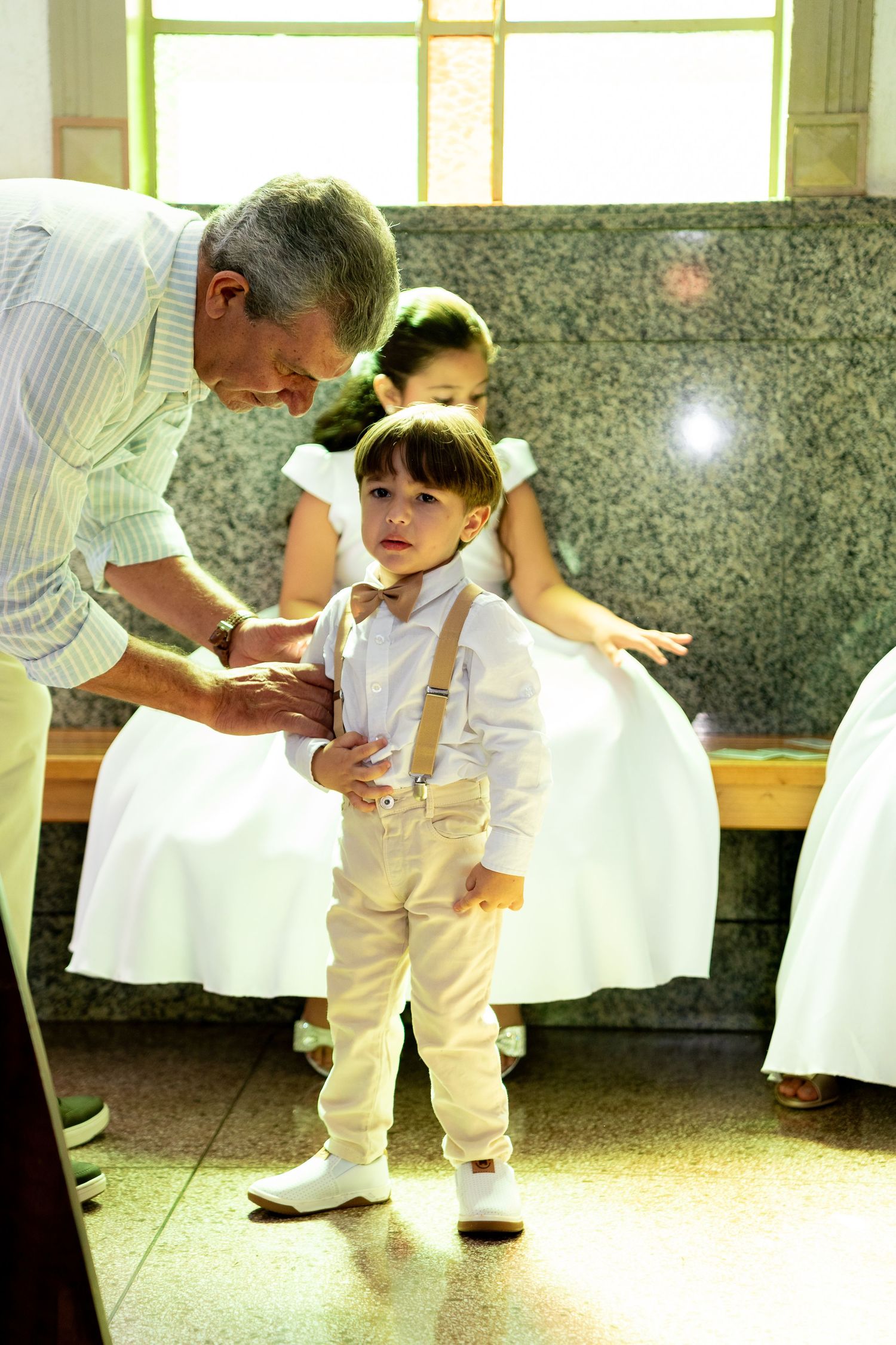 Young boy in suspenders and bow tie stands with an older man and girl in white dress at a formal event.