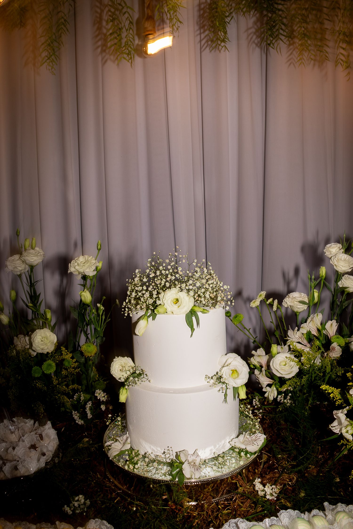 Elegant two-tier white wedding cake adorned with baby's breath and white roses, surrounded by floral arrangements.