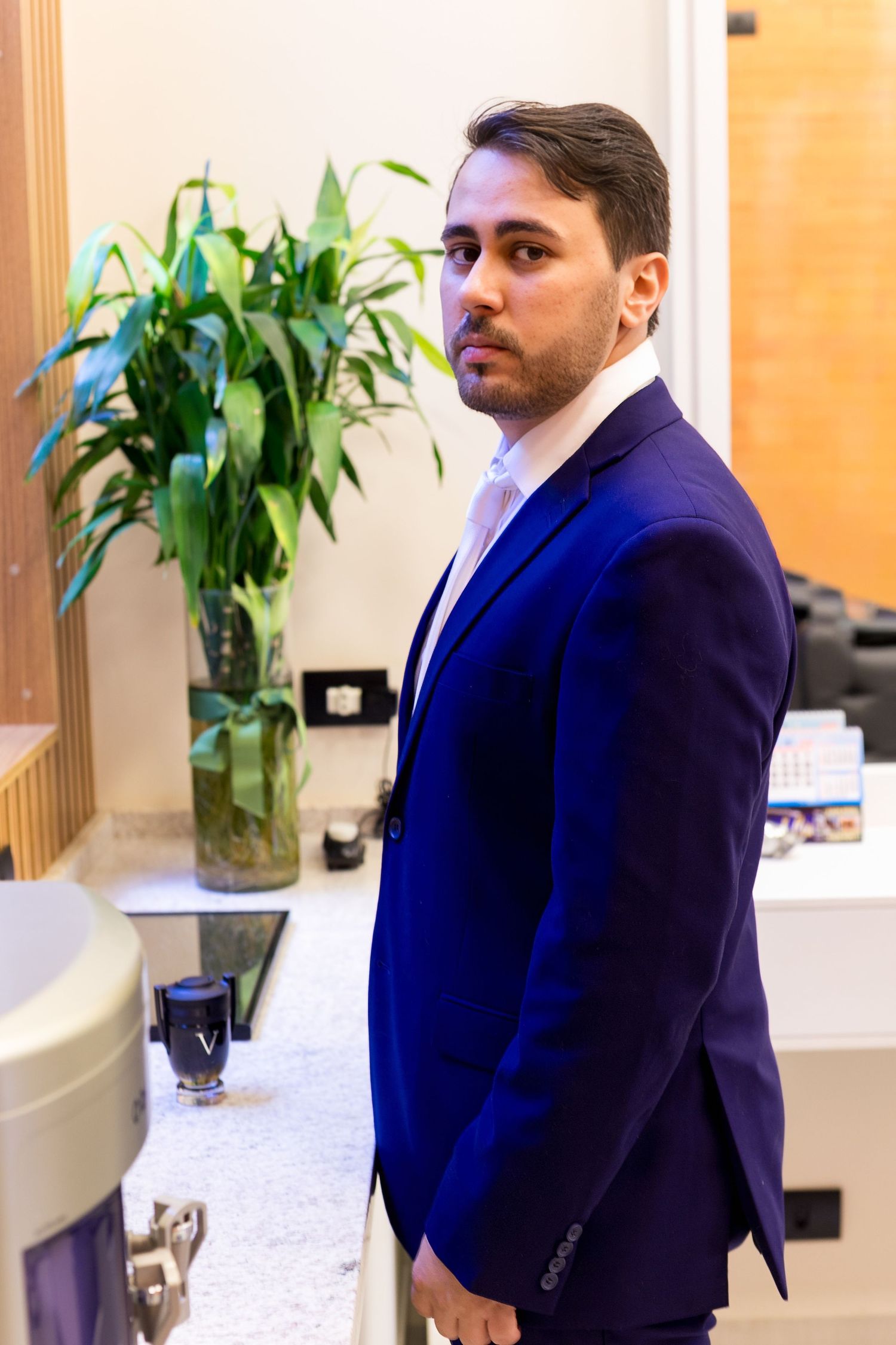Young man in blue suit standing near bathroom counter with green plant in background.
