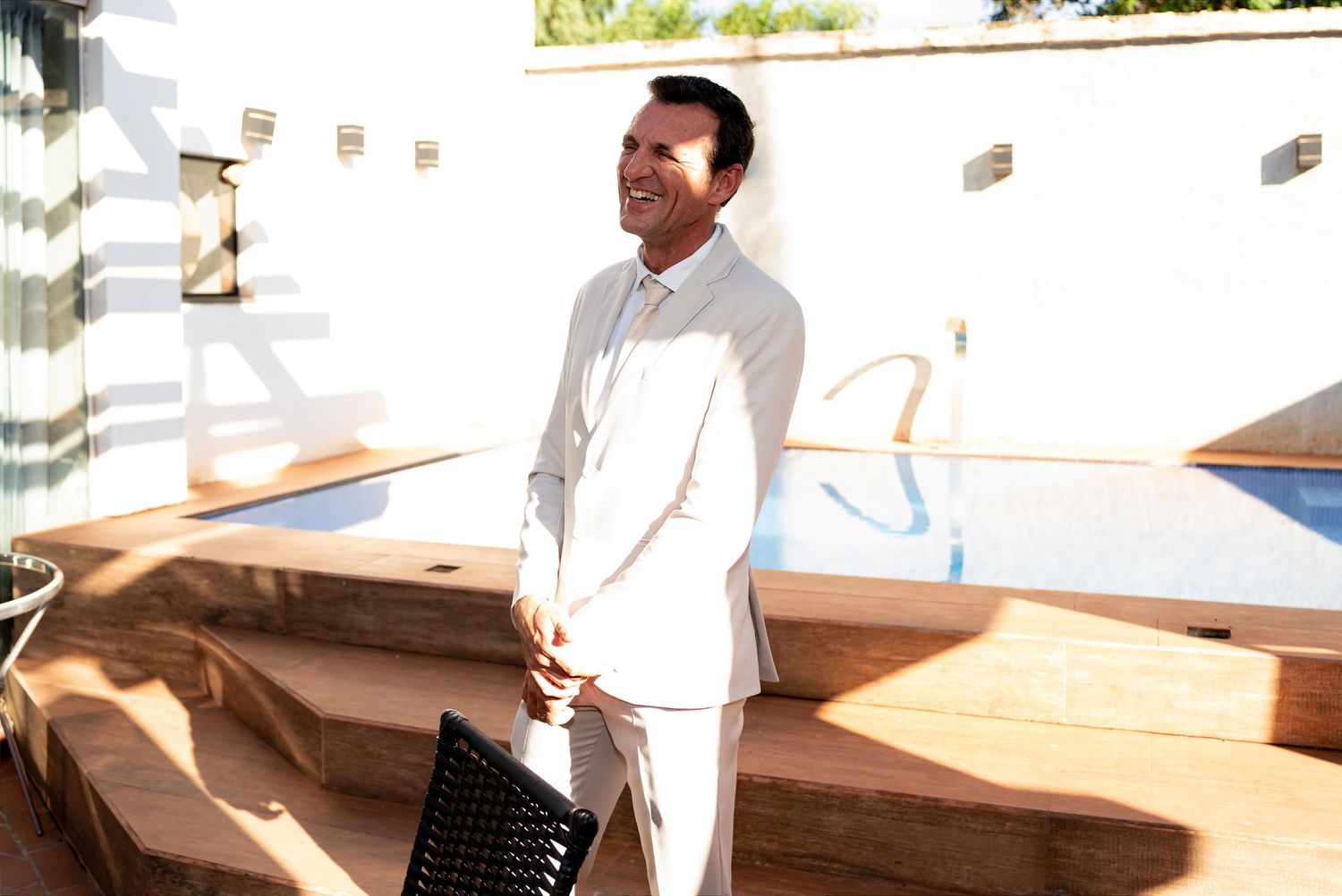 Man in white suit smiling outdoors by a pool on a sunny day.