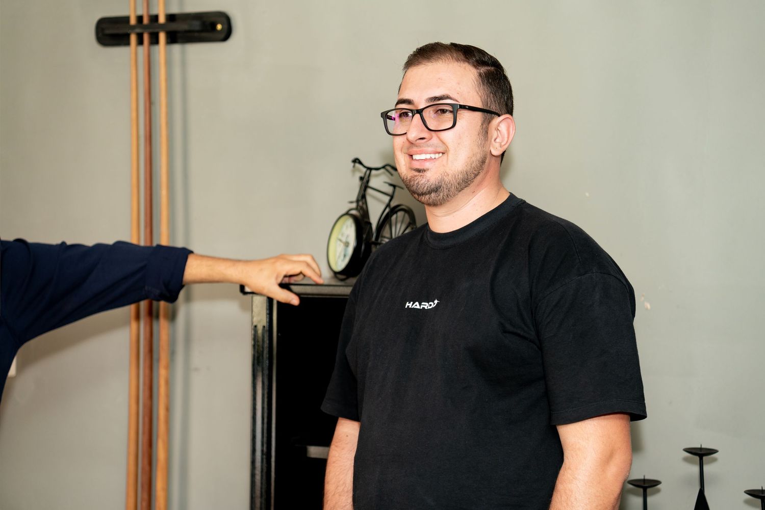 A smiling man wearing black glasses and a black t-shirt stands indoors while someone points at him.