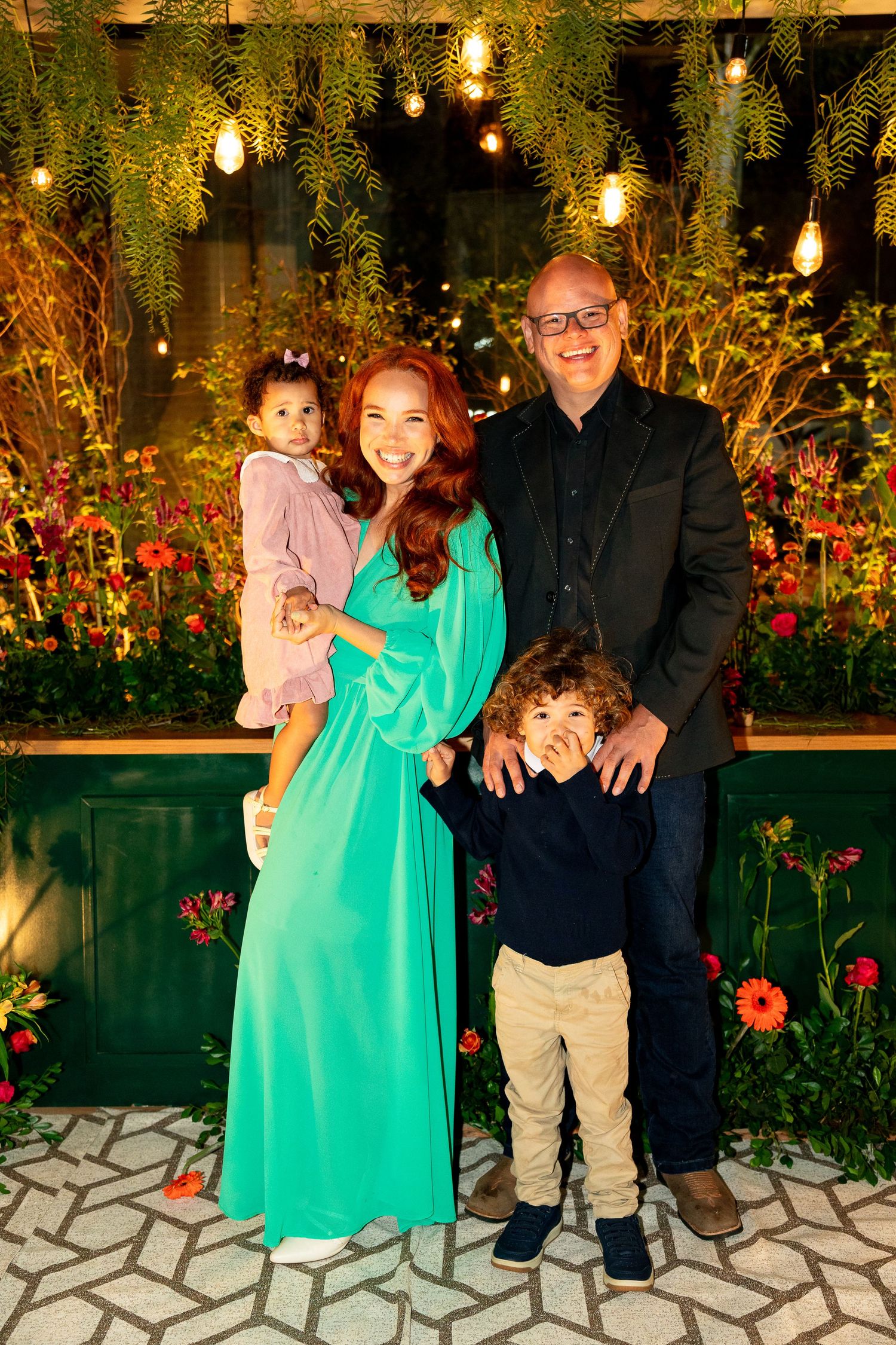 Family of four posing outdoors at night with string lights and lush greenery, woman in teal dress holding toddler.