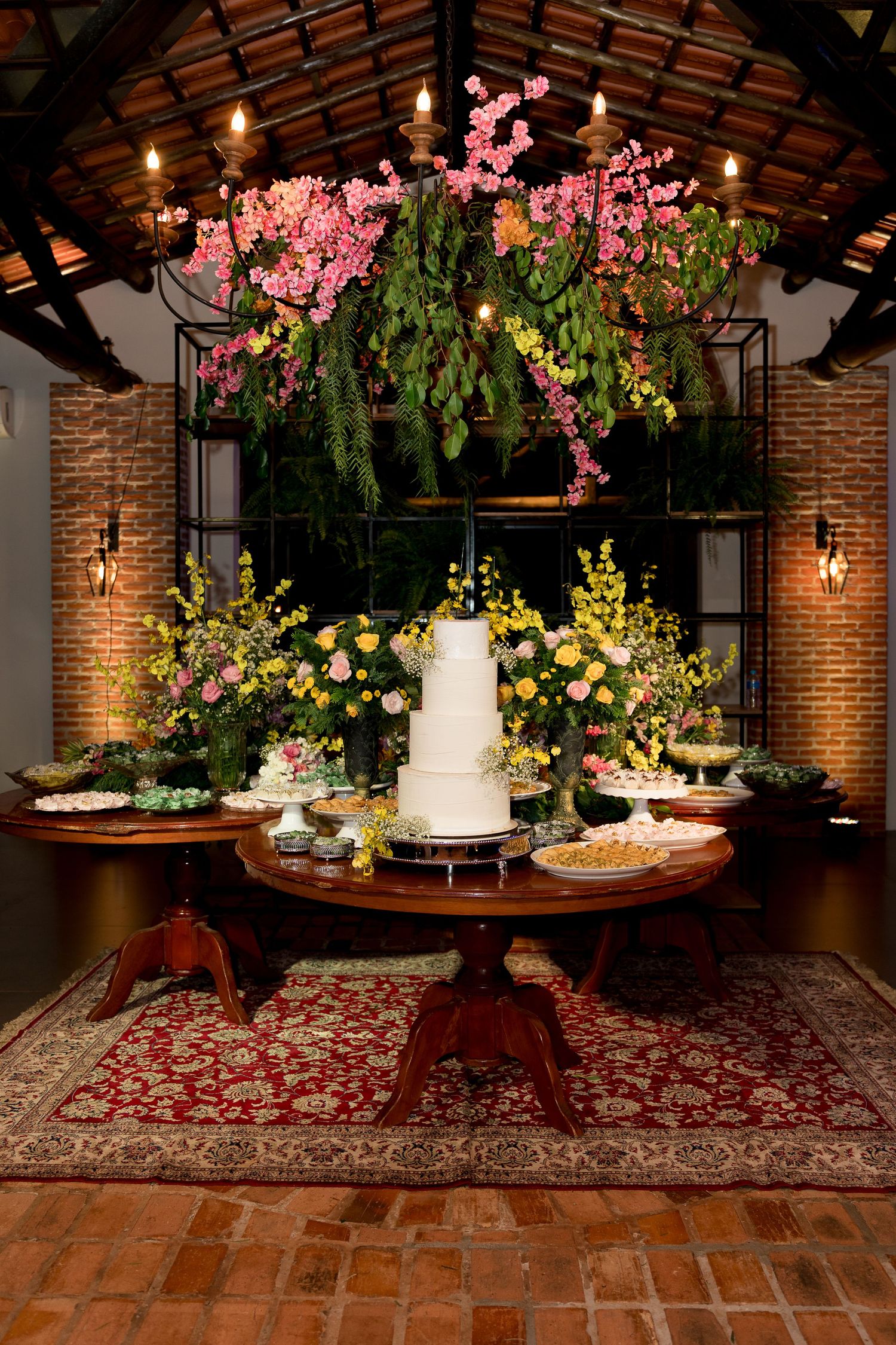 Elegant wedding dessert table with white tiered cake, floral arrangements, and hanging pink blooms in a rustic brick venue.