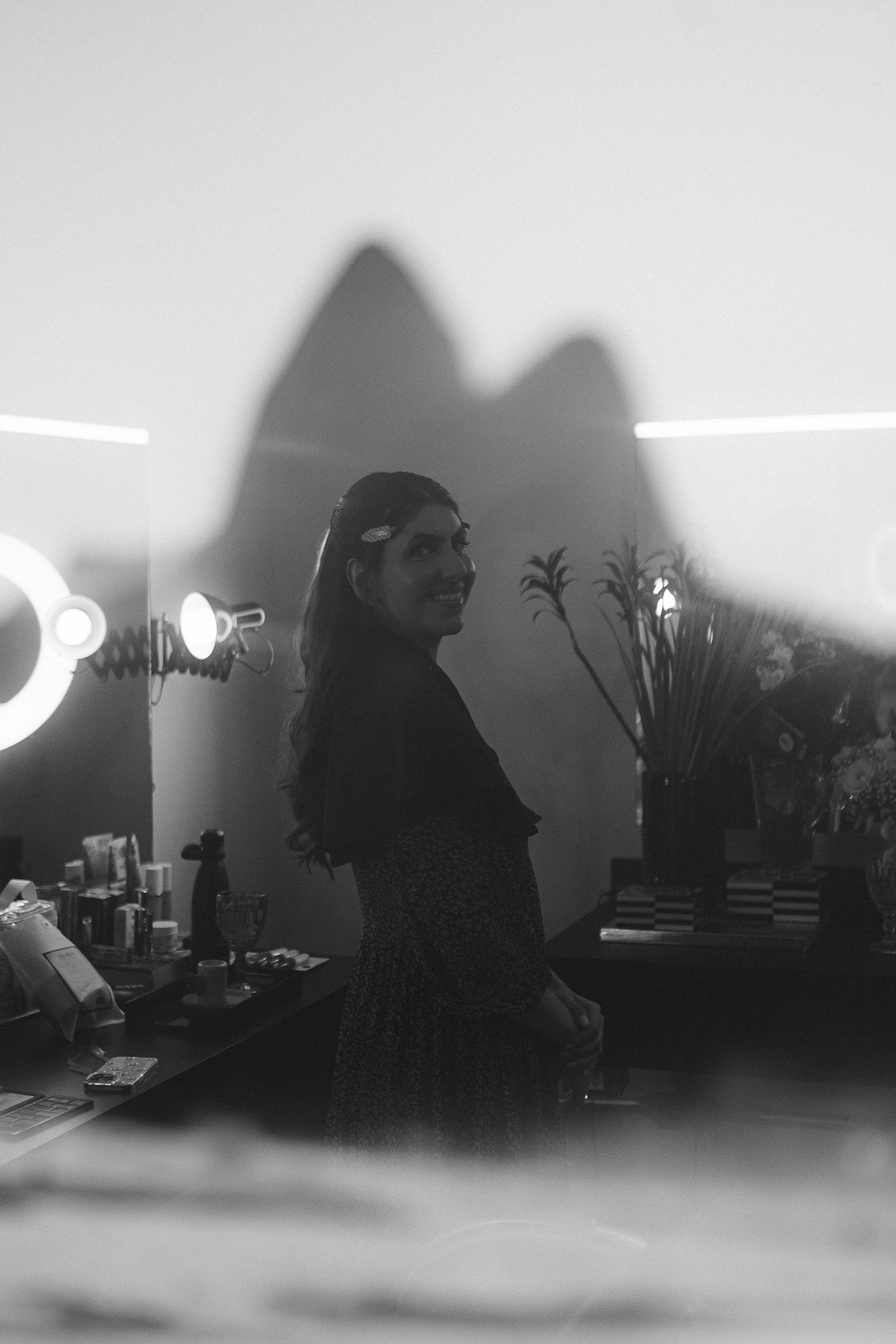 Black and white photo of a woman smiling in a dressing room with vanity lights and makeup table.