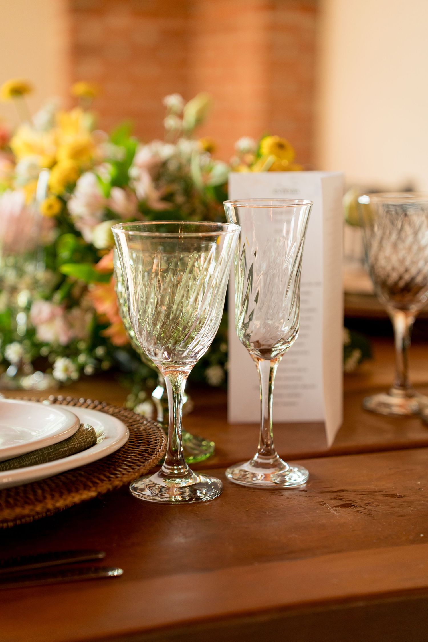 Elegant crystal wine glasses on a wooden dining table with floral centerpiece and woven charger plates.
