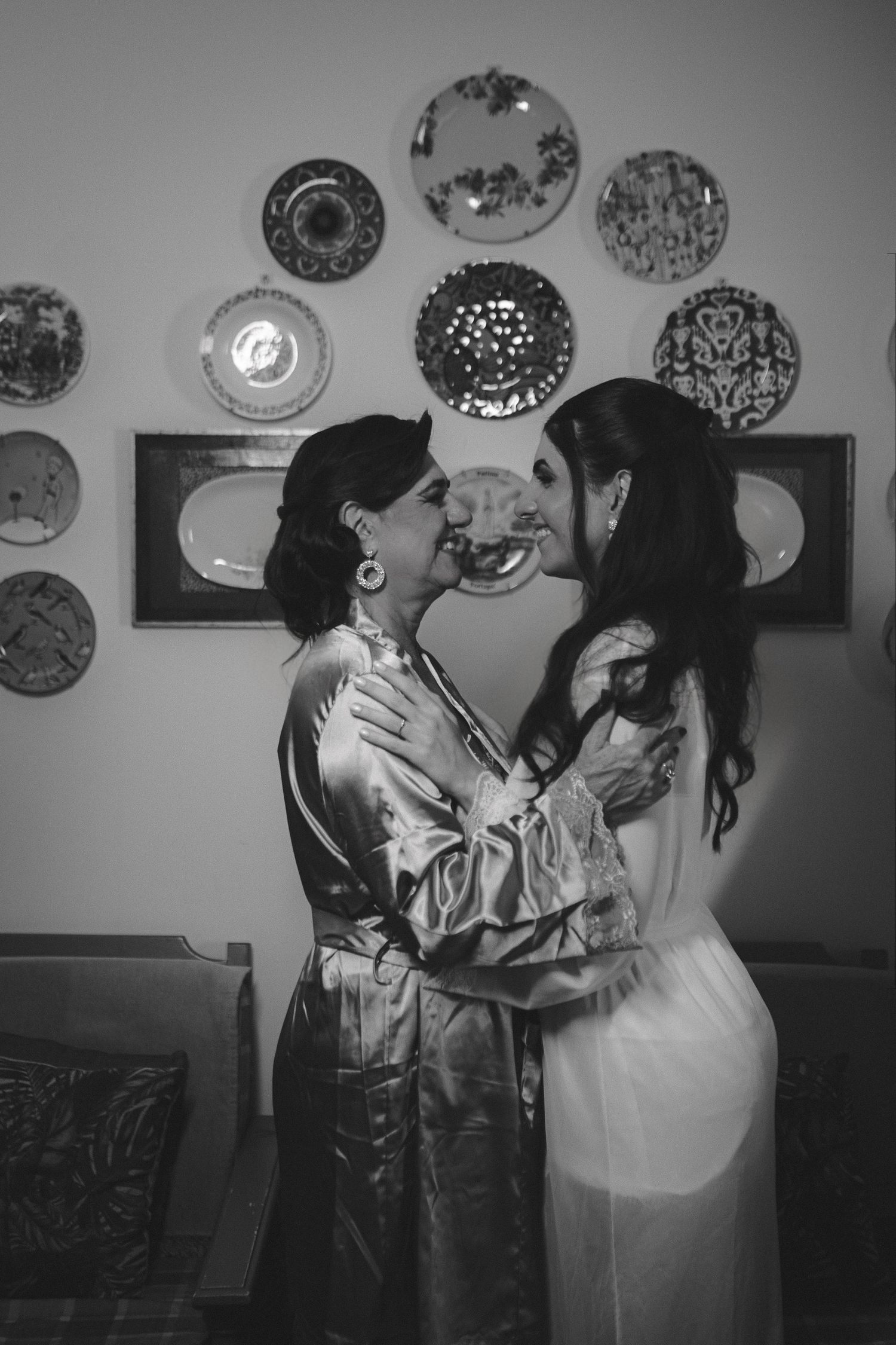 Black and white photo of two women in elegant attire embracing and kissing, with decorative plates displayed on the wall behind them.