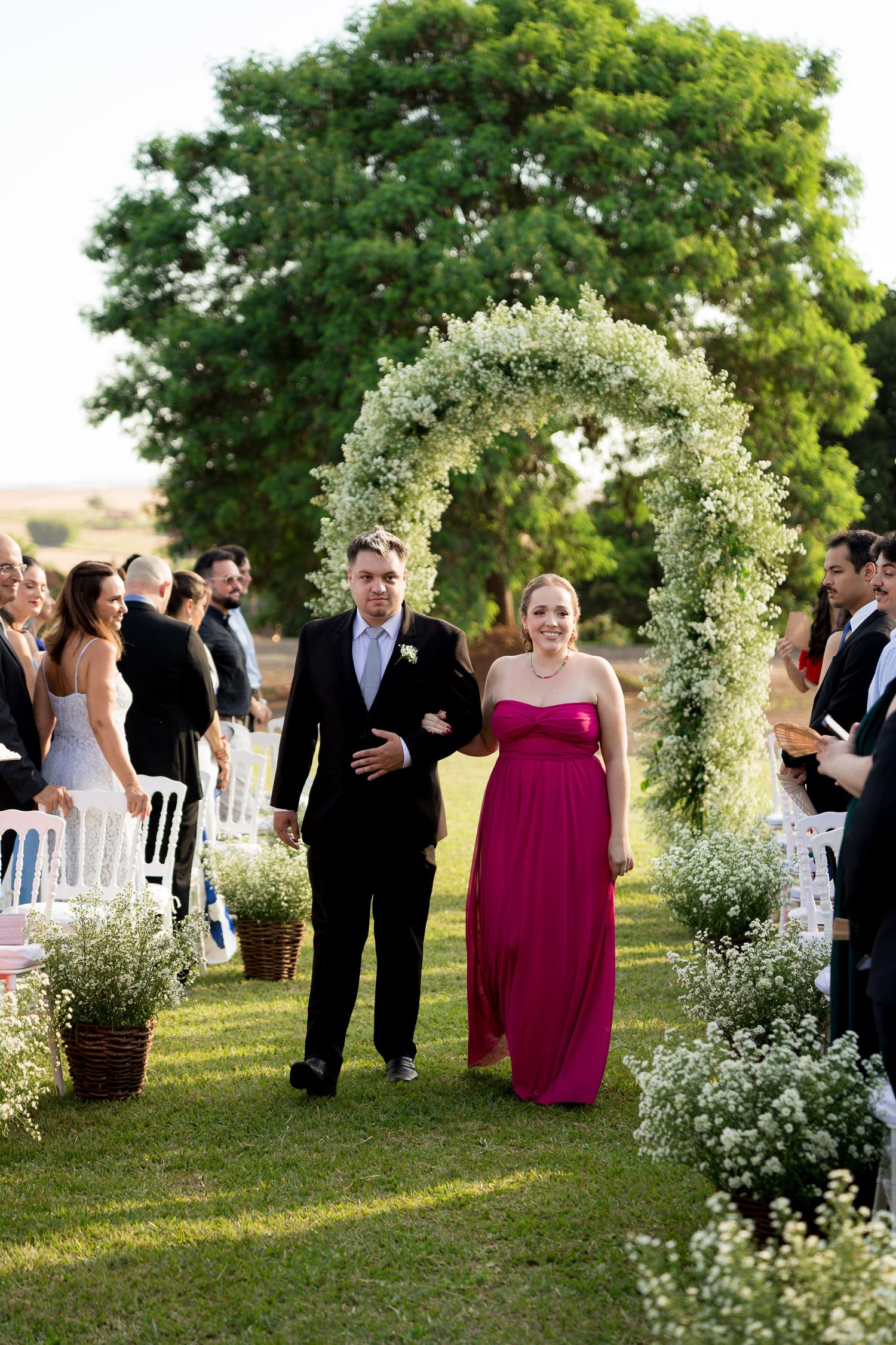 Couple walks down outdoor wedding aisle under floral arch, woman in pink gown, man in suit, white chairs and guests.