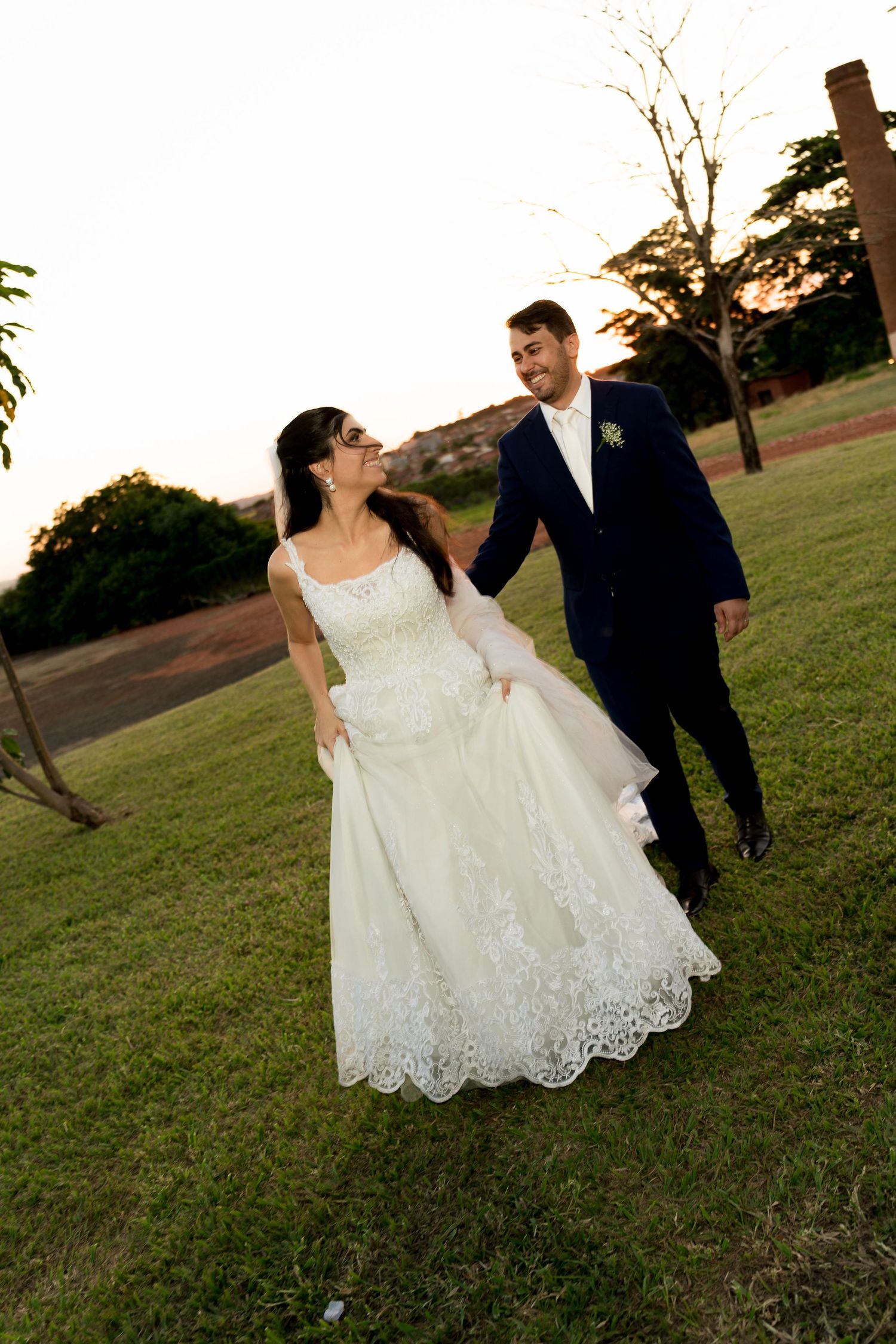 A bride in a white lace gown and groom in a navy suit walk hand-in-hand on a green lawn at sunset.