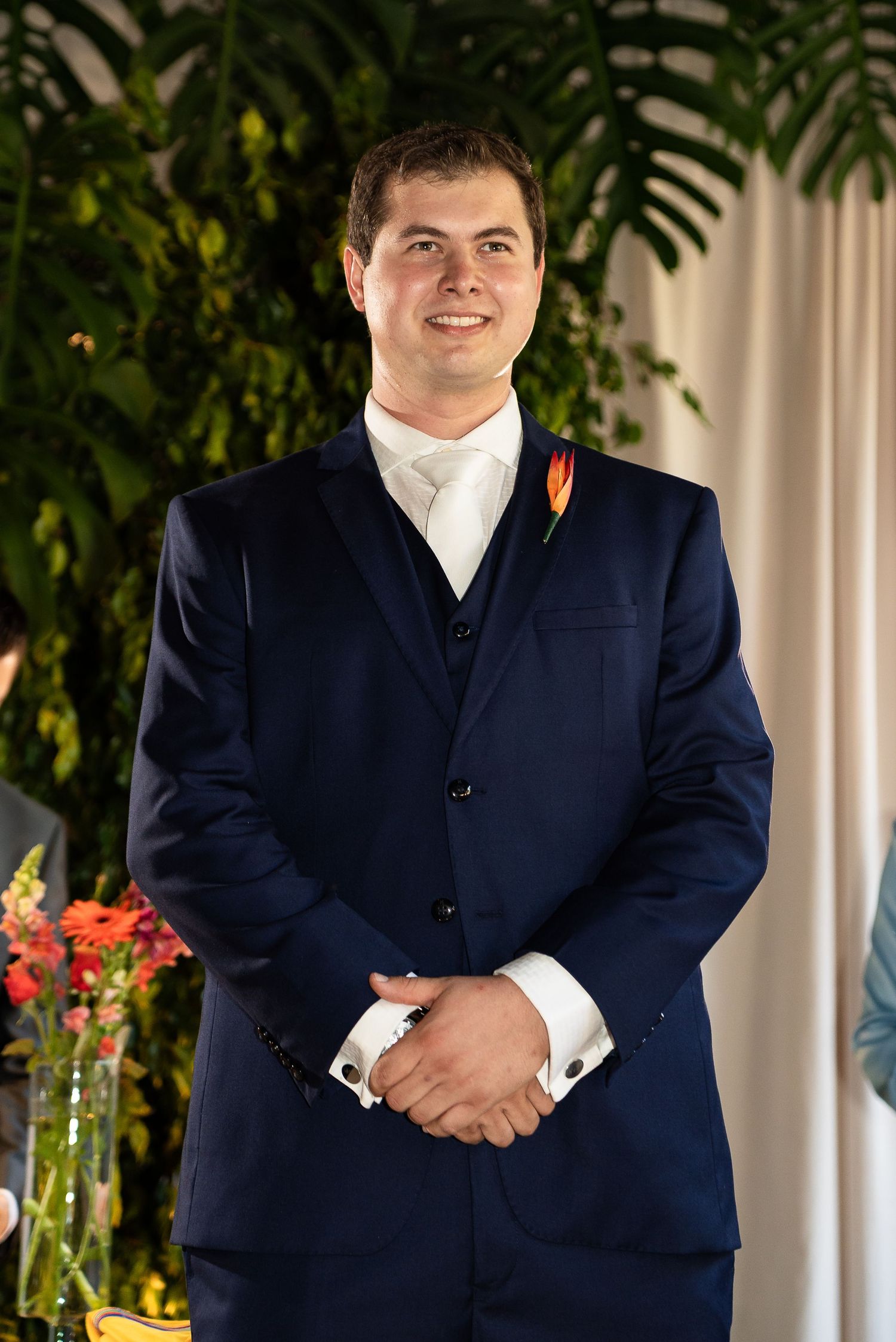 Smiling groom in navy blue suit with orange boutonniere stands at wedding altar with tropical greenery backdrop.