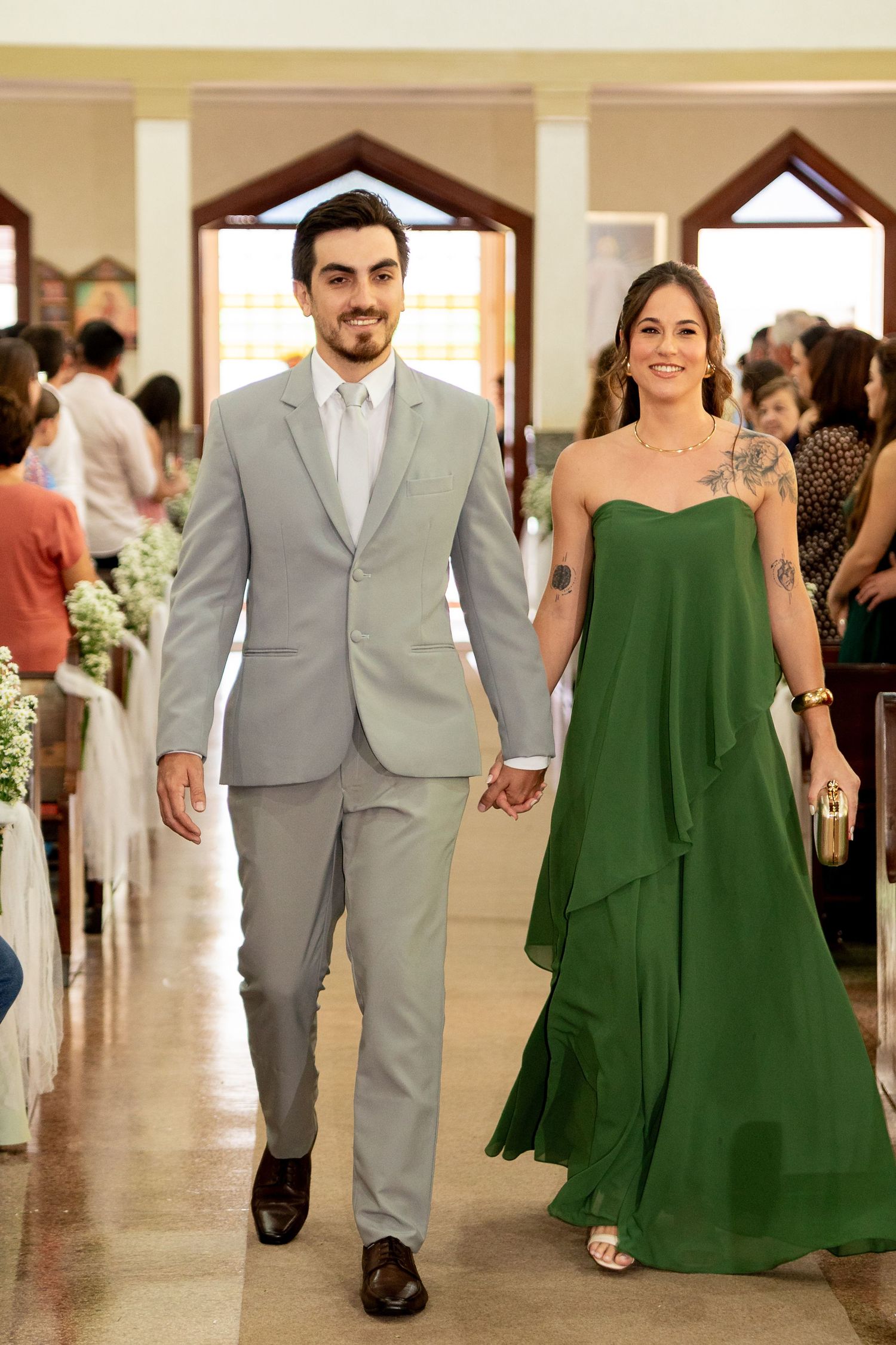 Couple walking down church aisle, man in gray suit, woman in strapless green gown, holding hands at wedding ceremony.