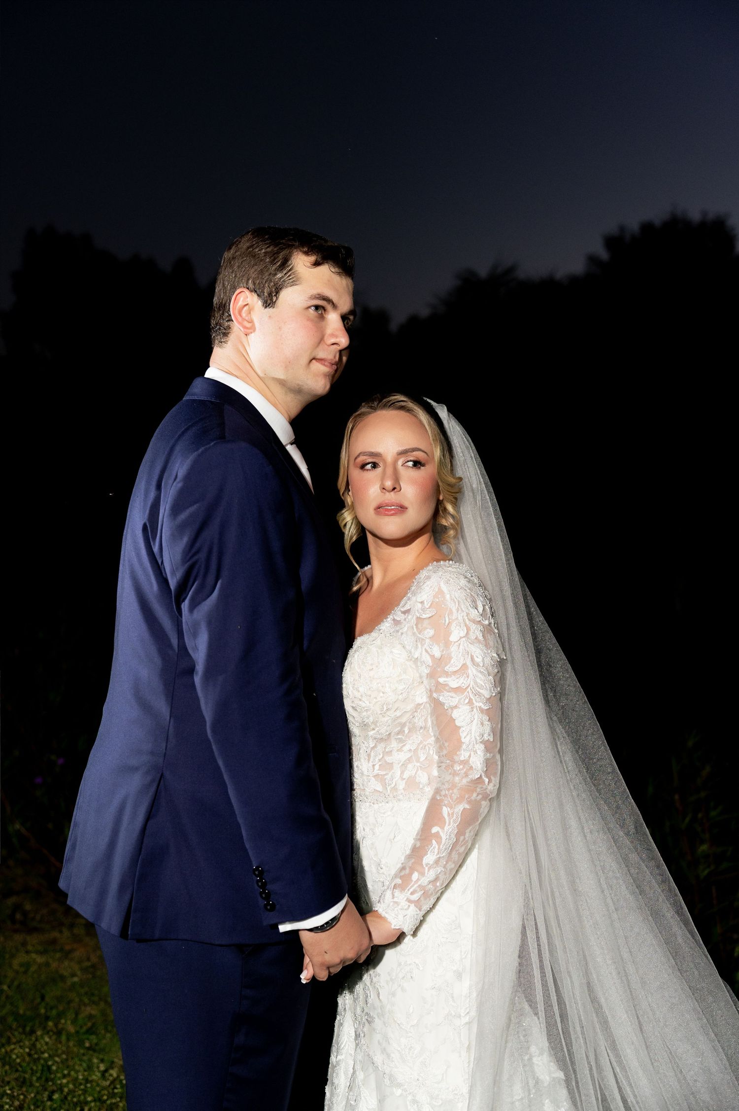 Bride and groom hold hands in elegant wedding attire, posing outdoors at night under a dark sky.