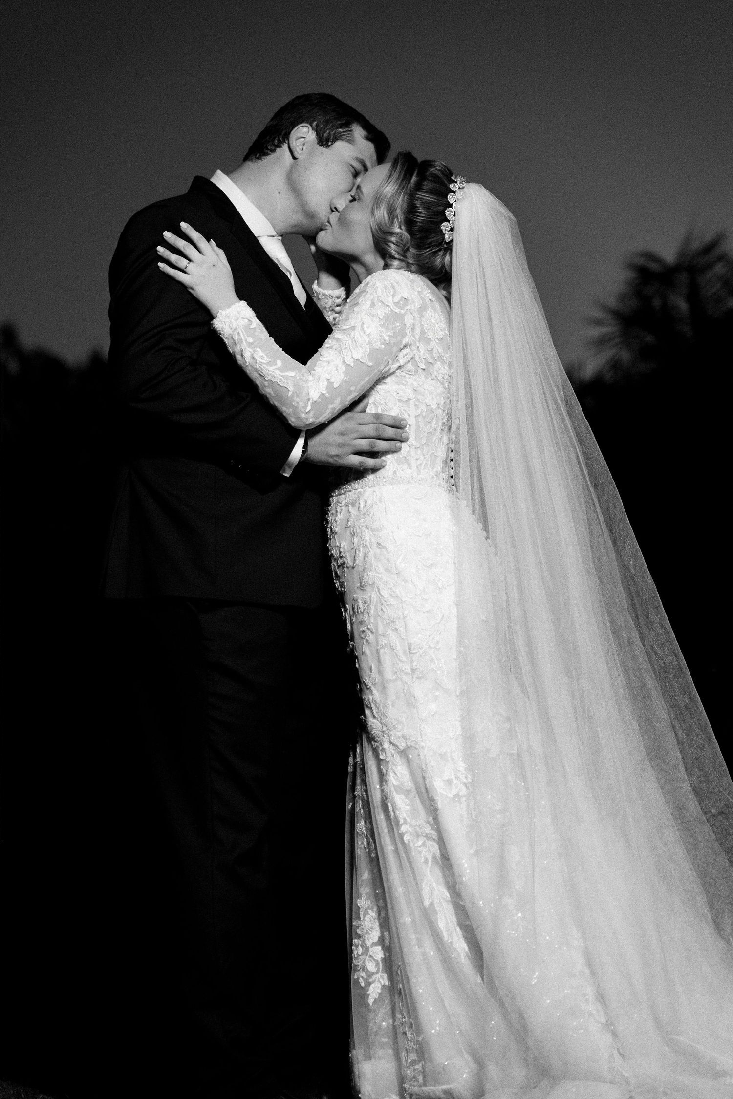 Black and white photo of a bride and groom sharing a romantic kiss at dusk, bride wearing lace gown and veil.