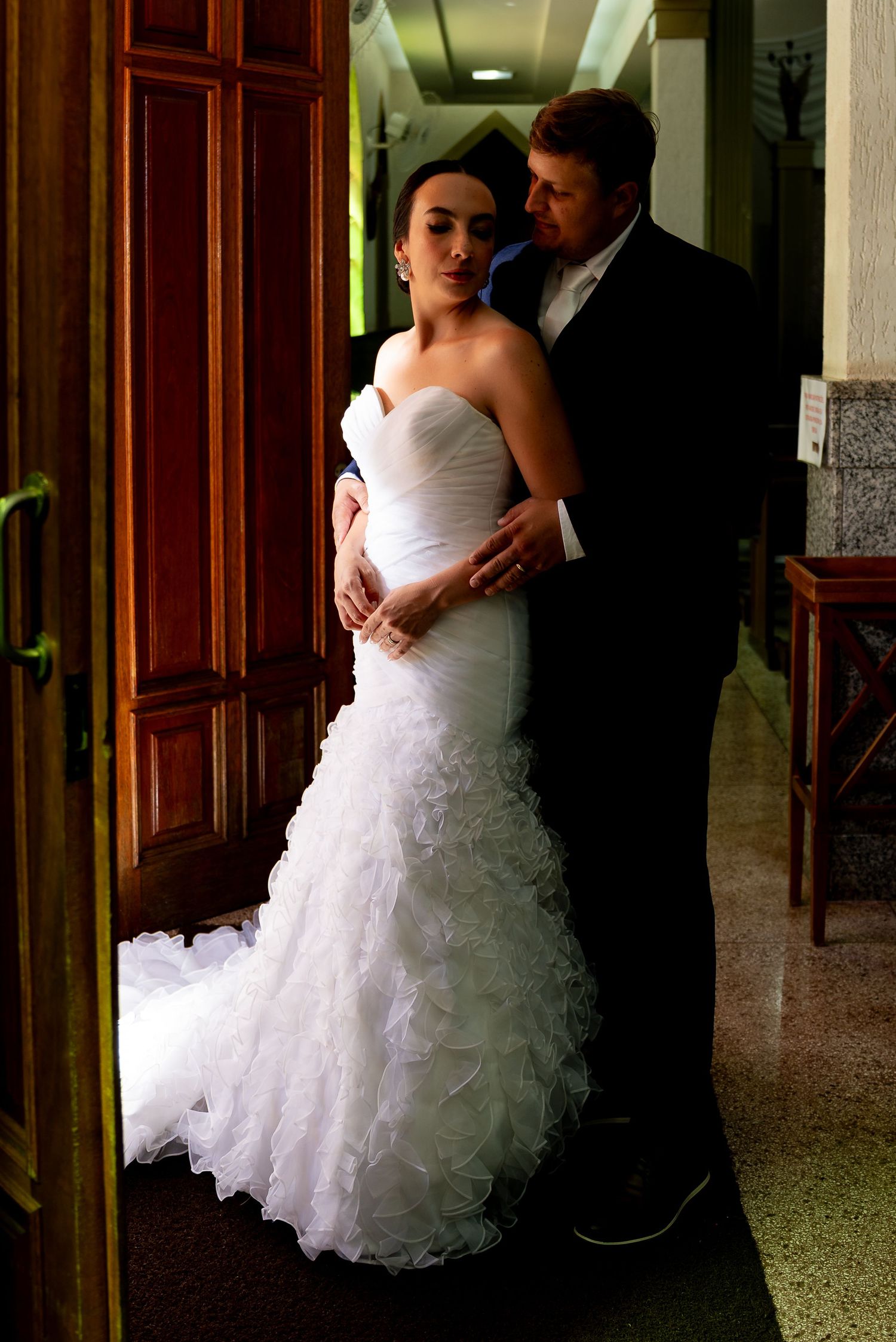 Elegant bride in white mermaid gown with ruffled train stands with groom in black suit in a doorway.