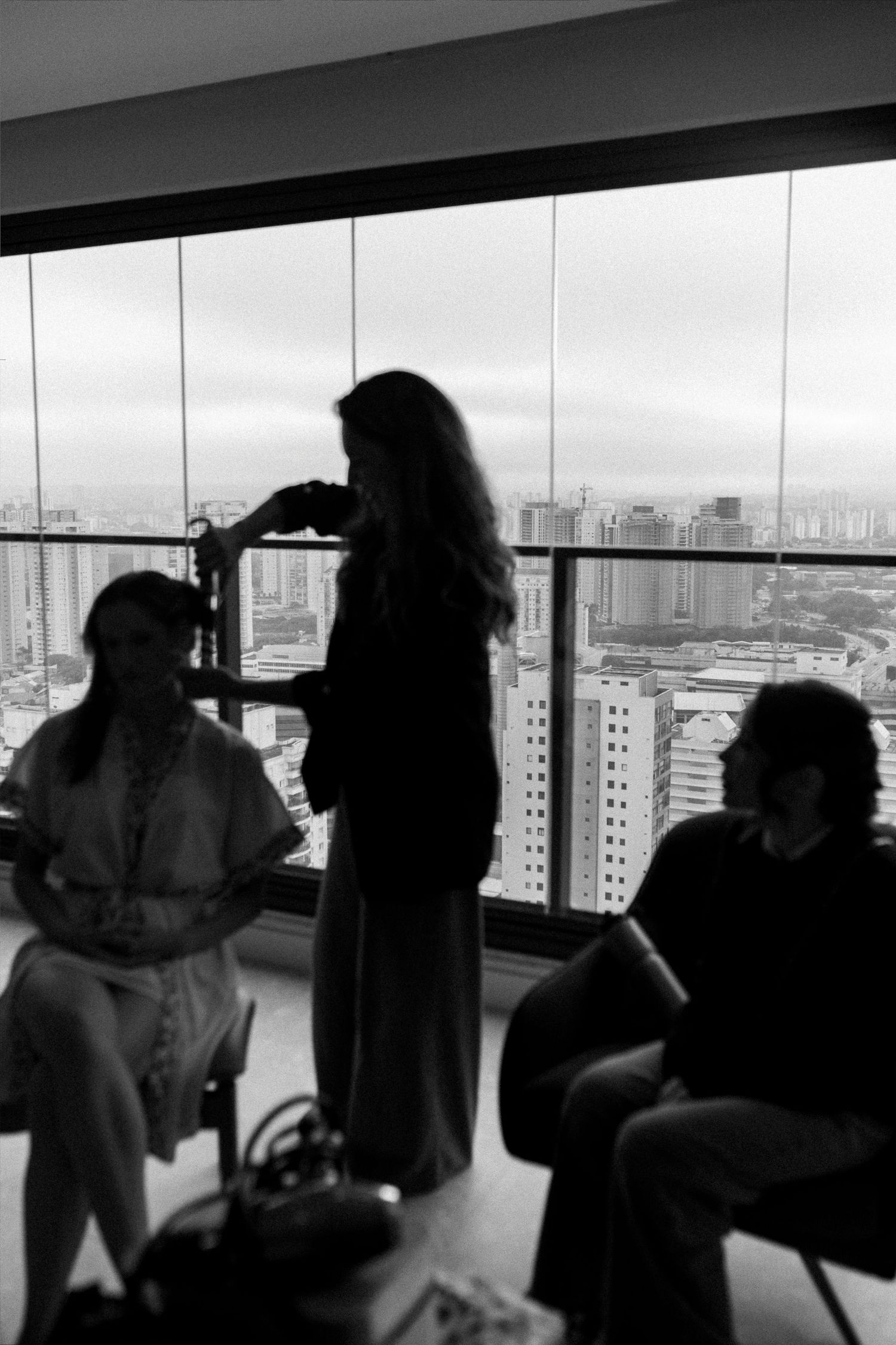 Black and white photo of three people in a high-rise with city skyline view, one woman standing by floor-to-ceiling windows.