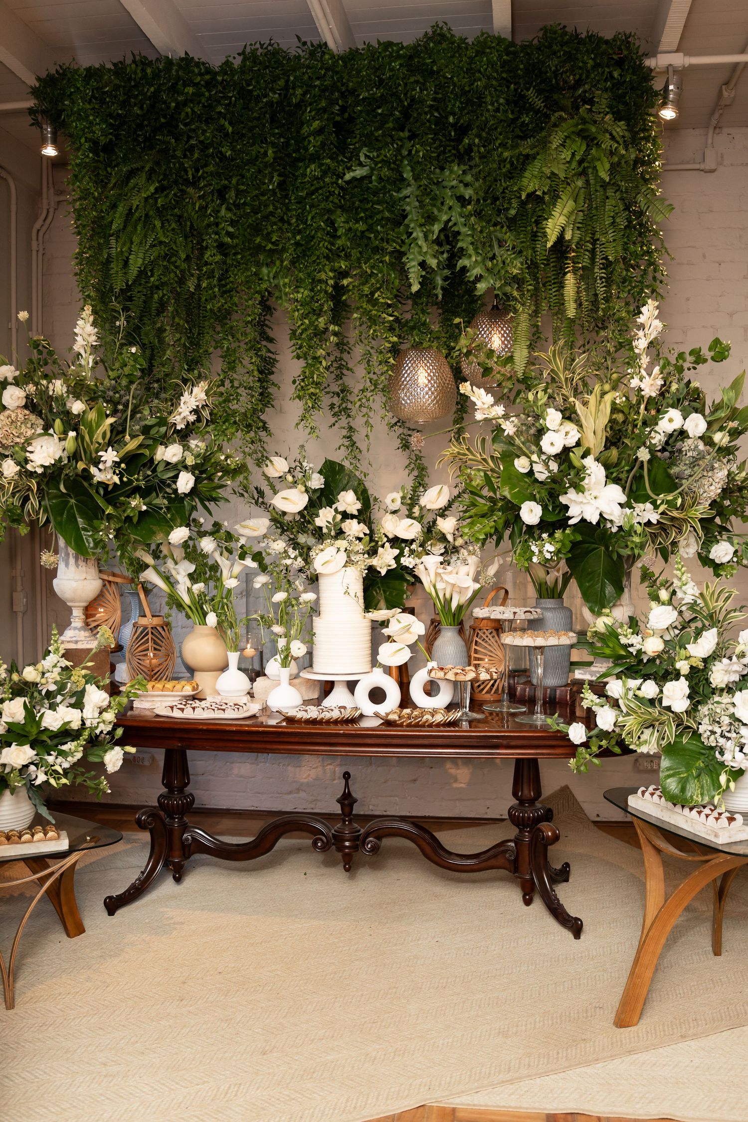 Elegant wedding dessert table with white floral arrangements, greenery backdrop, and tiered cake on ornate wooden table.