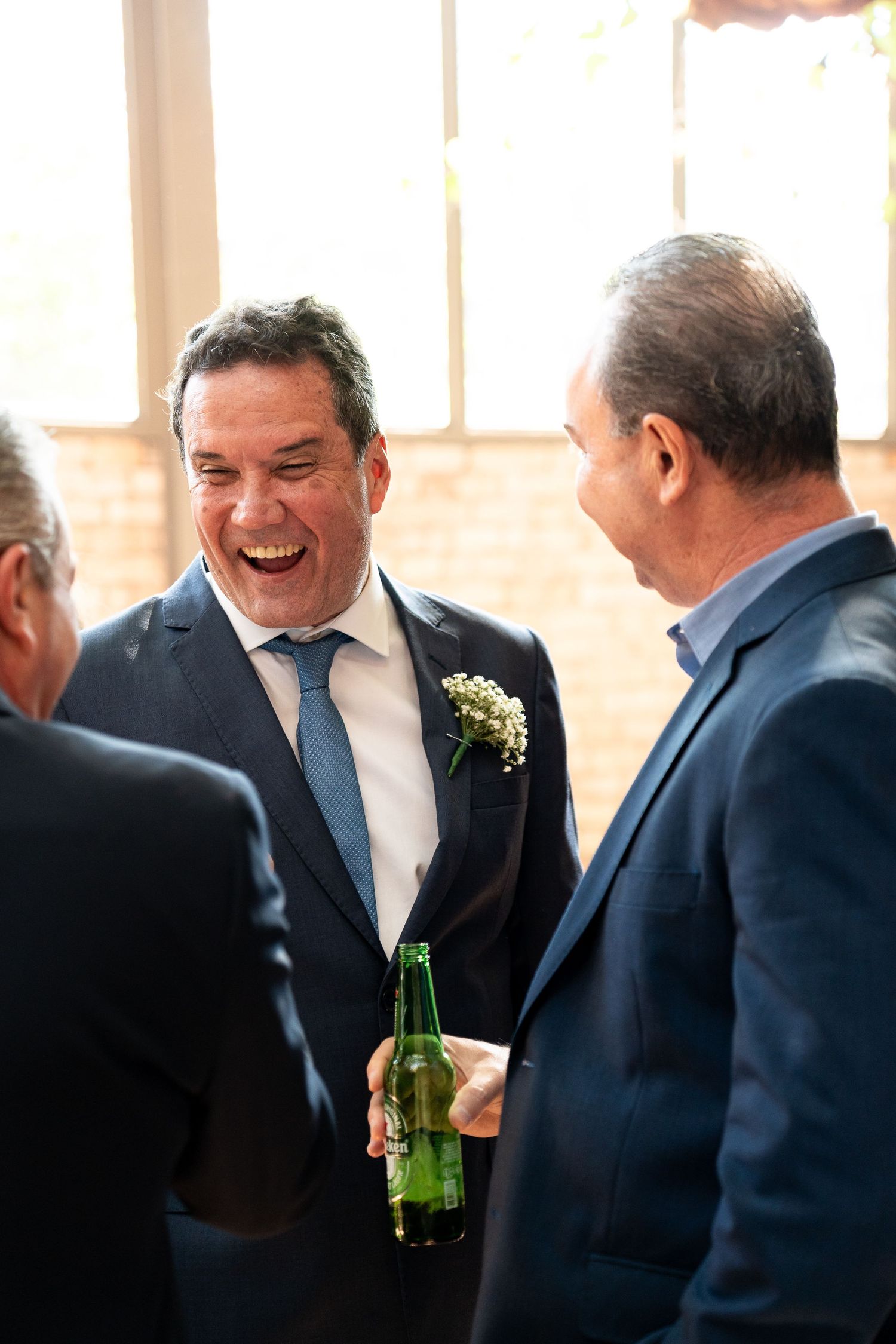 Man in navy suit with boutonniere laughing joyfully while holding a green beer bottle at a wedding reception.