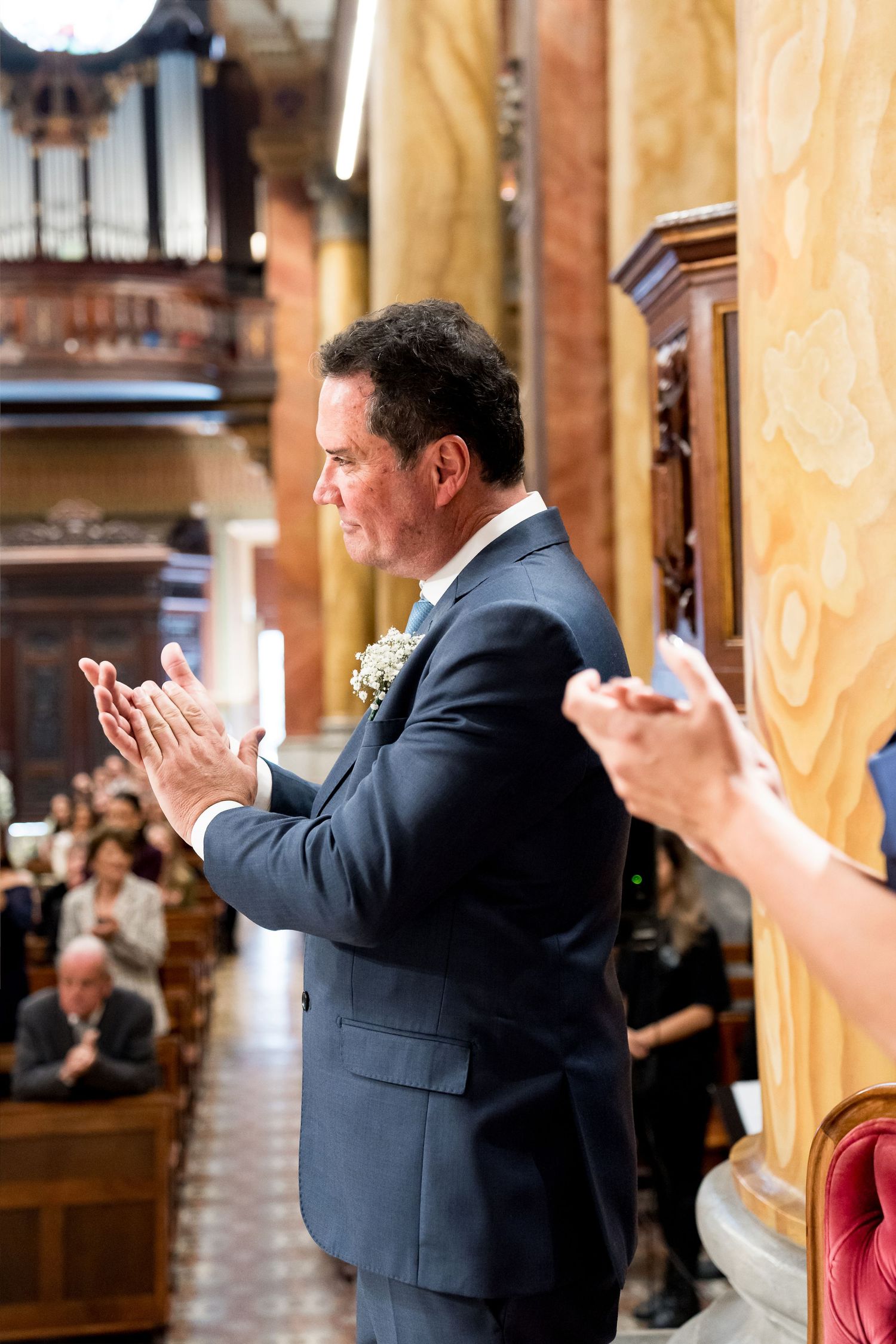 A man in a navy suit with a boutonniere claps at a church wedding ceremony, surrounded by seated guests.