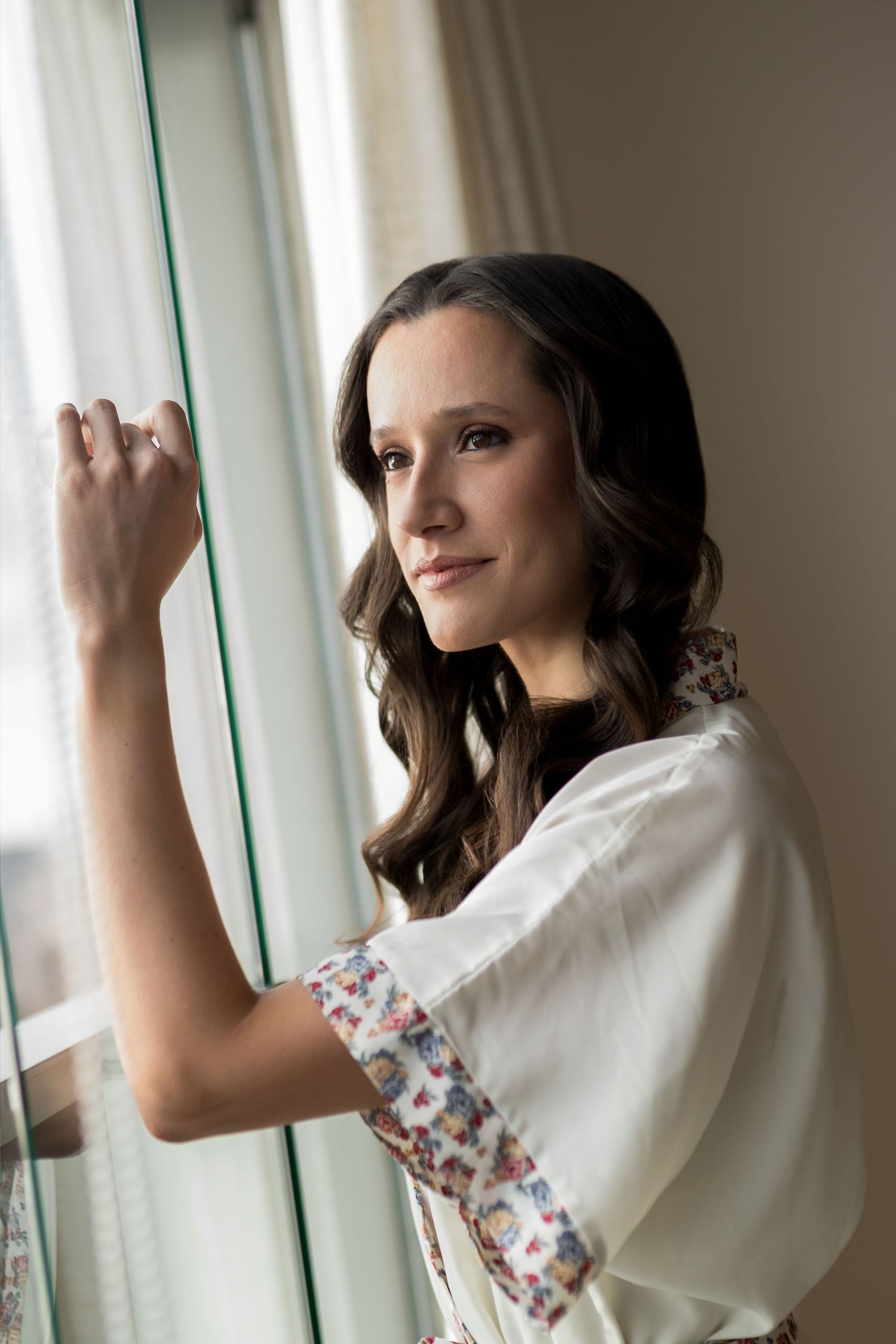 Young brunette woman in white floral robe gazing out a window with a gentle smile, soft natural light.