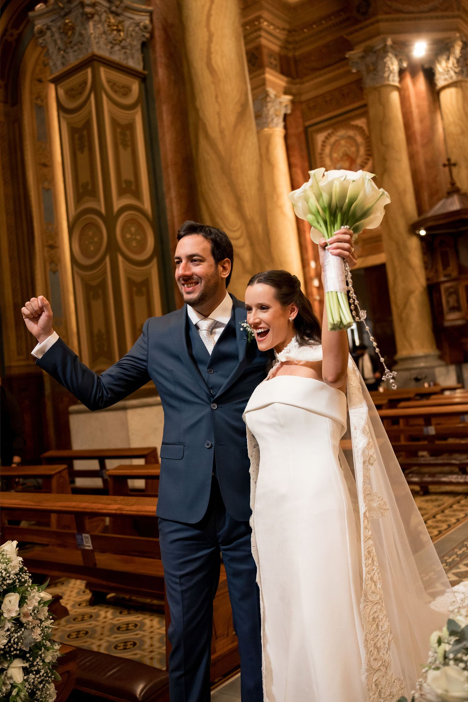 Joyful newlyweds celebrate in a church, bride raising bouquet and groom pumping fist in navy suit.