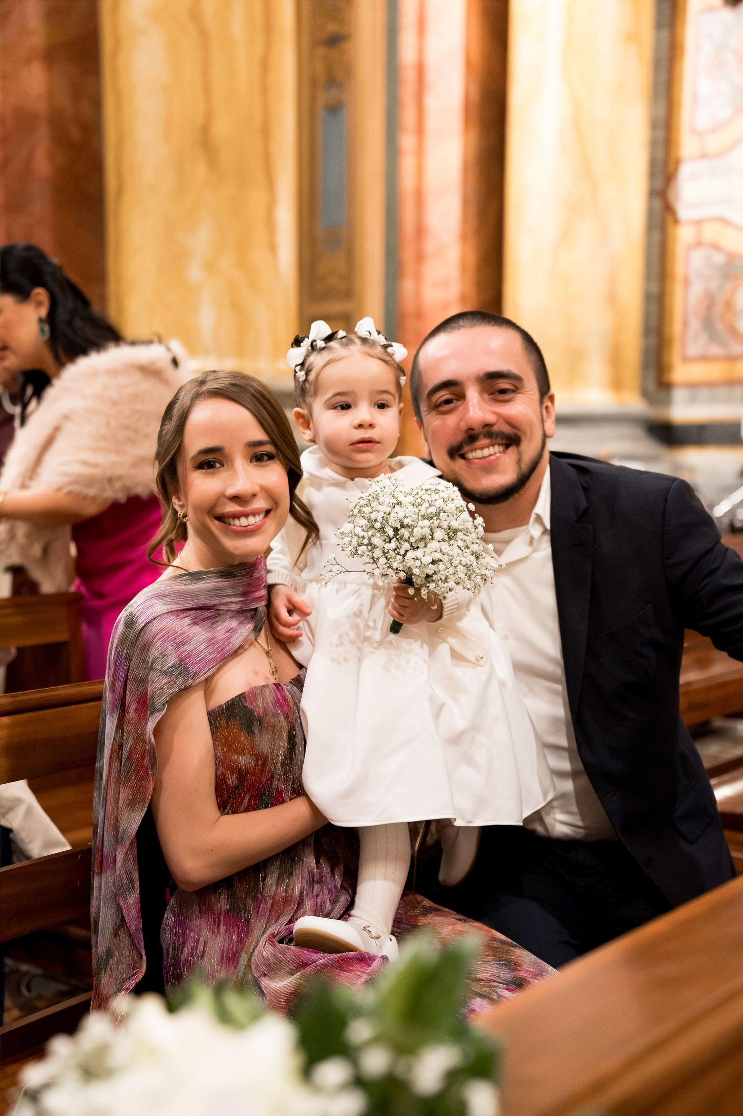 A smiling couple poses with their young flower girl holding a baby's breath bouquet inside an elegant church.