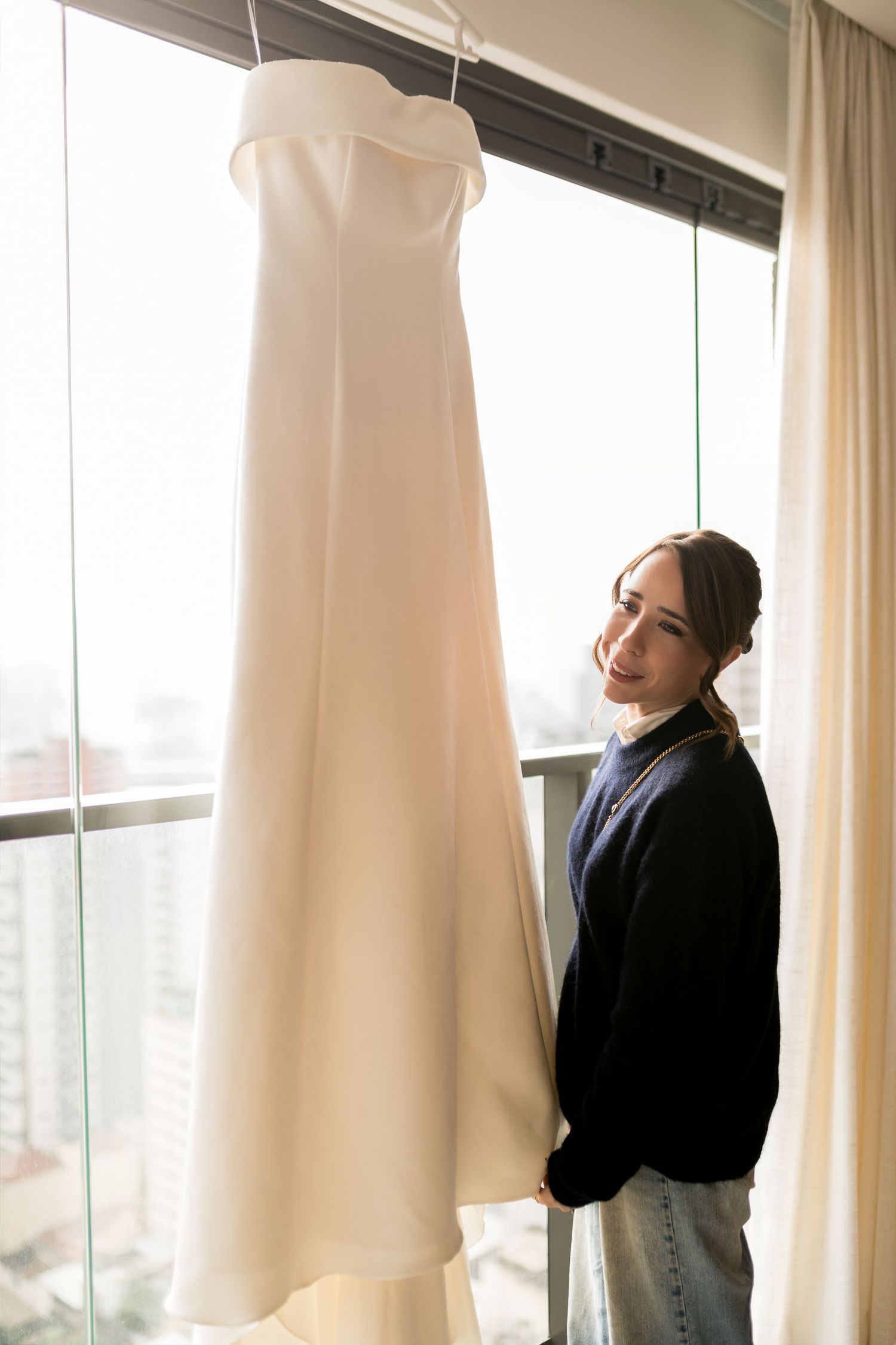 Smiling bride standing beside an elegant white wedding dress hanging by a bright window.