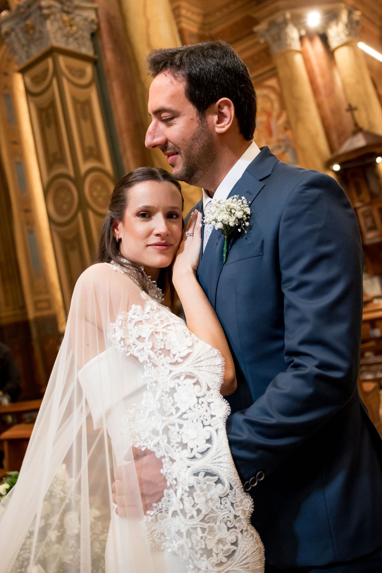 Bride in lace gown and veil embraces groom in navy suit with boutonniere inside ornate church.