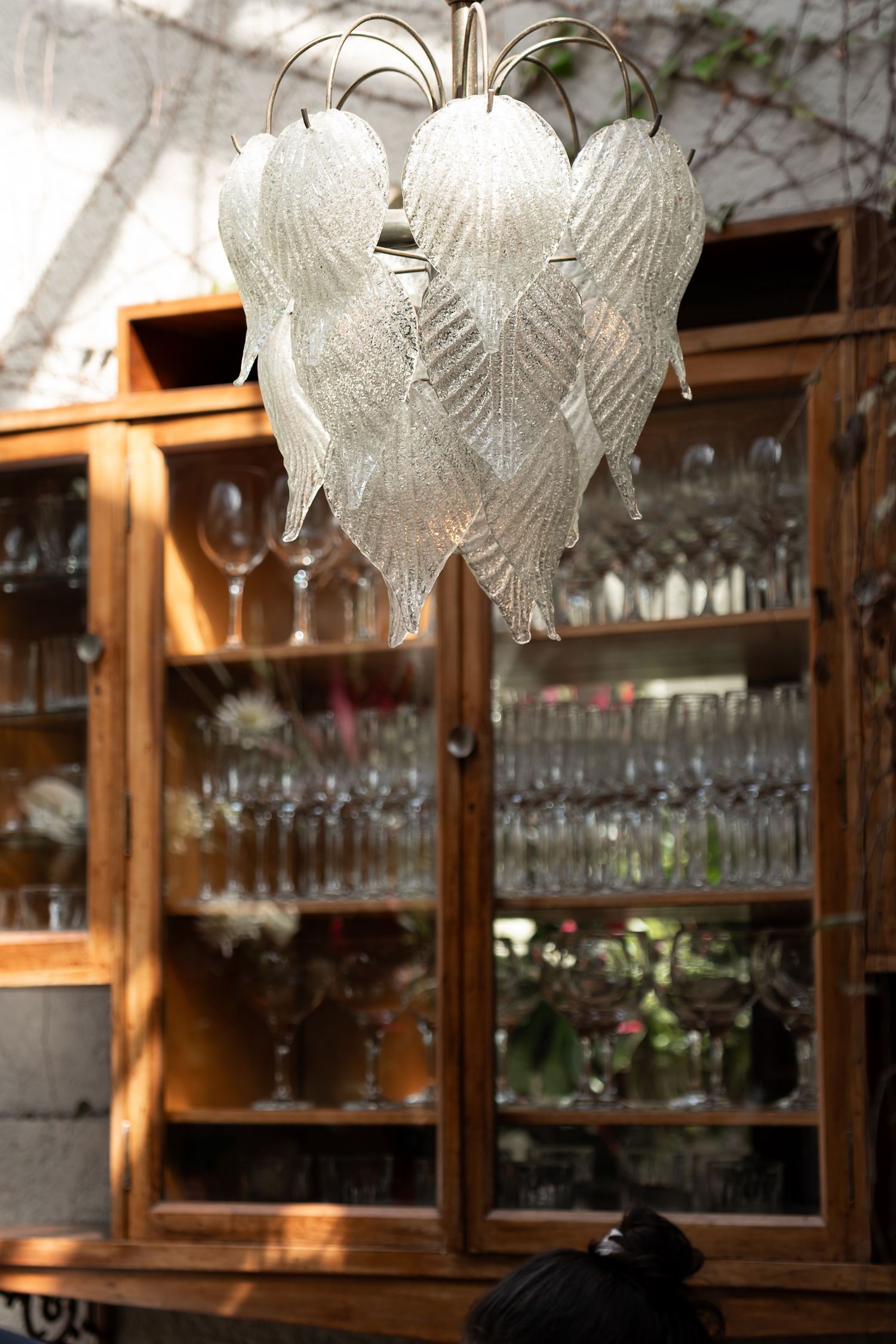 Elegant Murano glass leaf chandelier hanging in front of a wooden cabinet filled with crystal glassware.