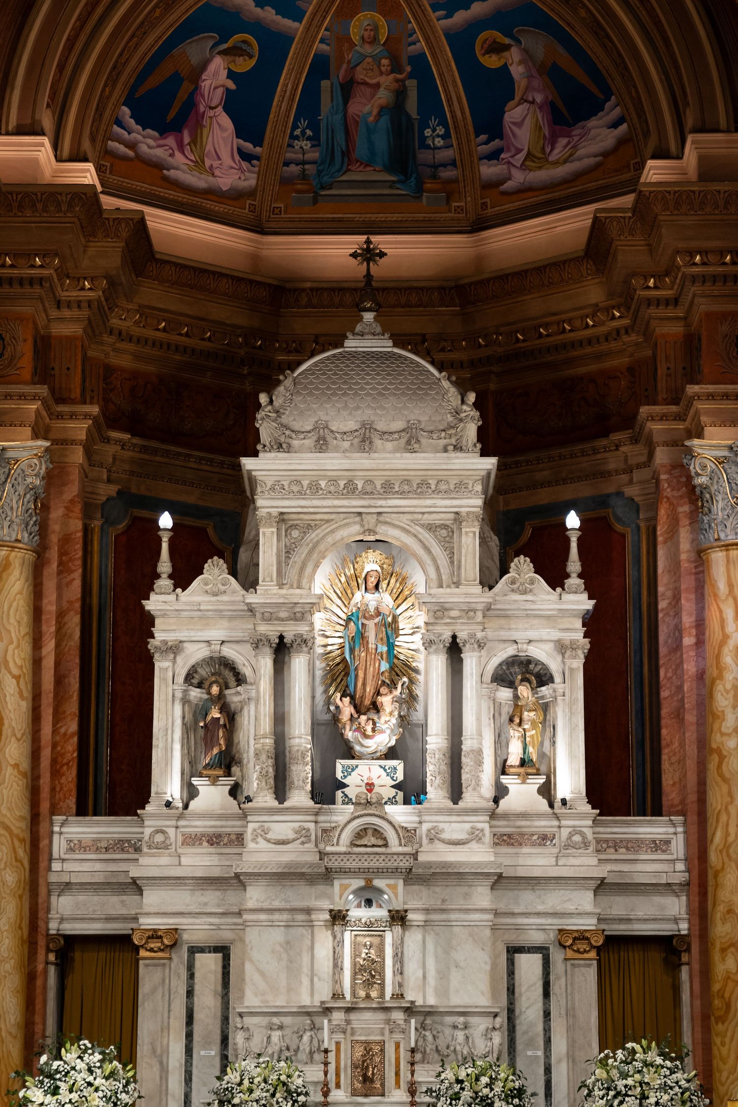 Ornate Catholic church altar with marble reredos, religious statues, candles, and colorful apse fresco above.