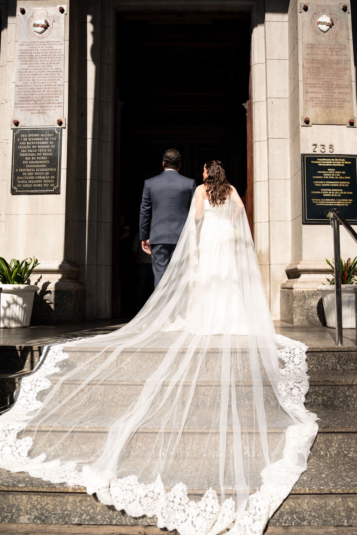 Bride in lace-trimmed cathedral veil and groom in navy suit entering church doors on wedding day.