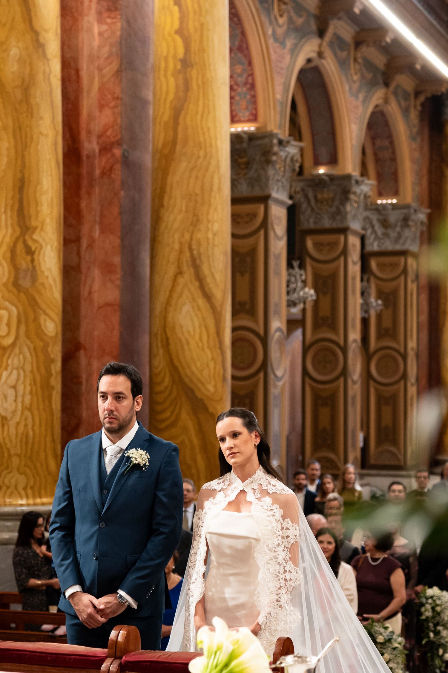 Bride in lace wedding gown and groom in blue suit standing at church altar during wedding ceremony.