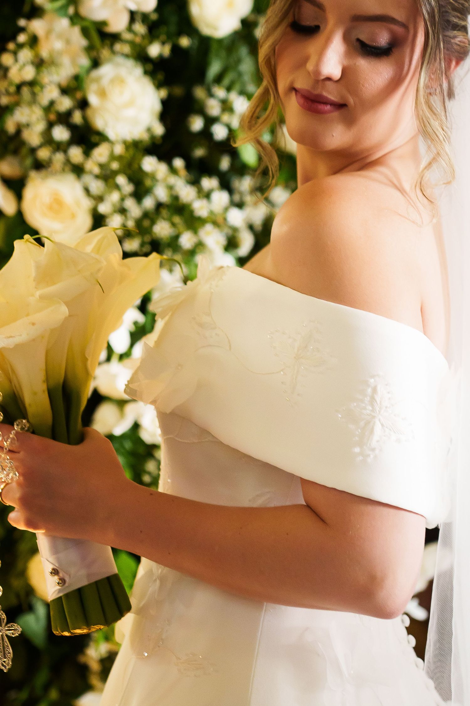 Elegant bride in off-shoulder white gown holding calla lily bouquet, standing before lush floral backdrop.