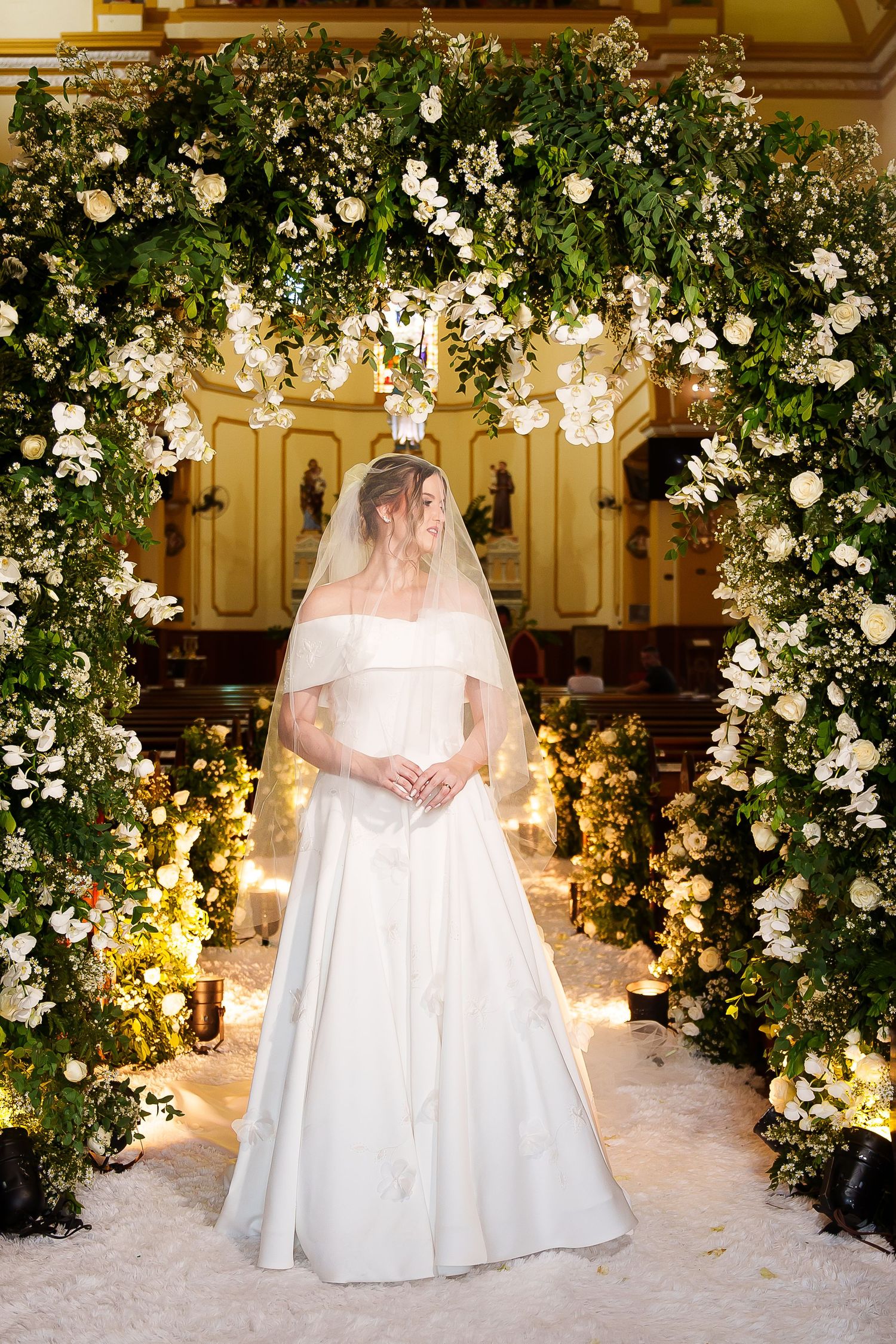 Bride in white off-shoulder gown and veil stands under lush floral arch with white roses in elegant church.