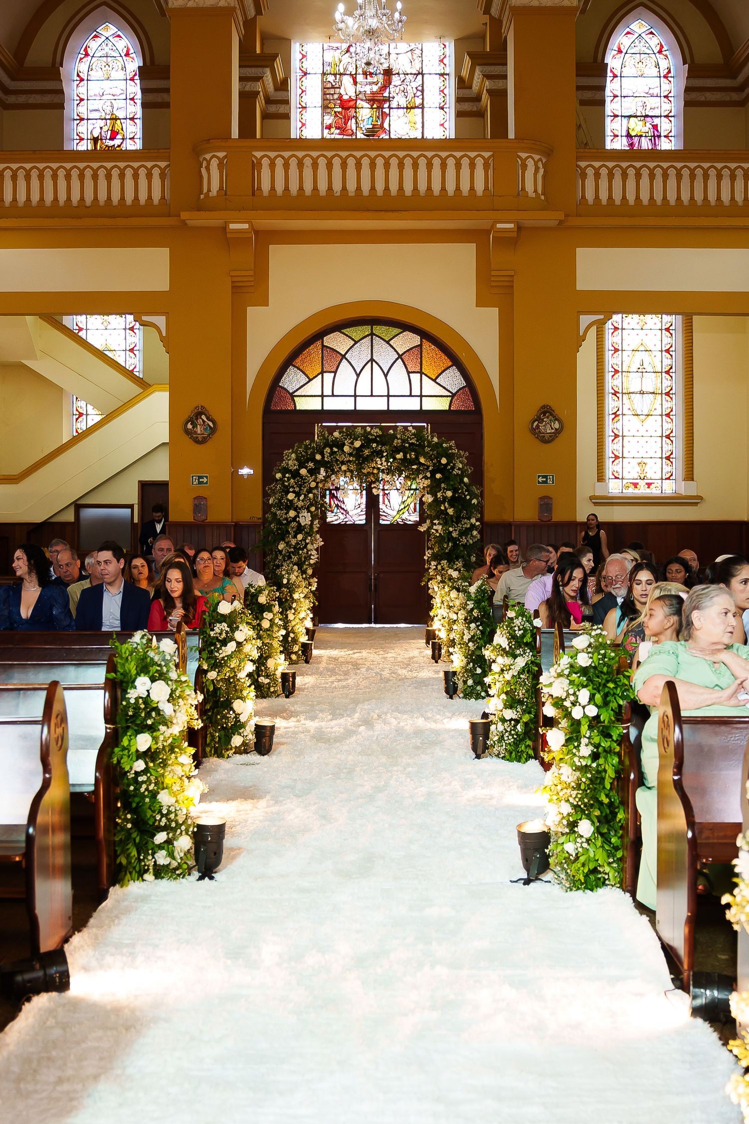 Elegant church wedding aisle with white carpet, lush greenery, floral arrangements, wooden pews, and stained glass windows.