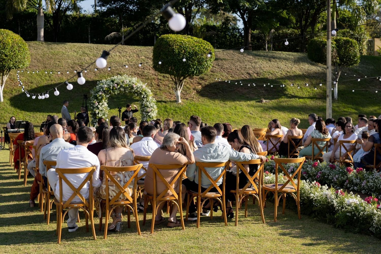 Guests seated in wooden cross-back chairs at an outdoor wedding ceremony on a lush green lawn.