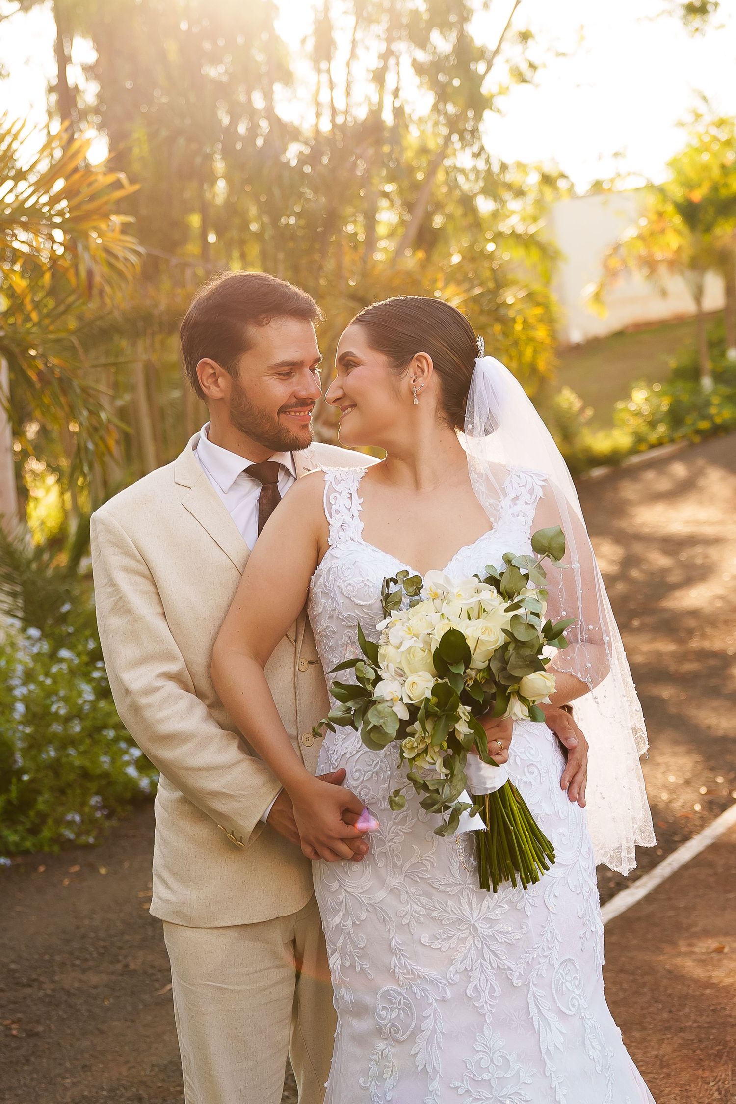Bride and groom share a romantic moment outdoors, bride holding white bouquet in lace gown with veil, groom in tan suit.