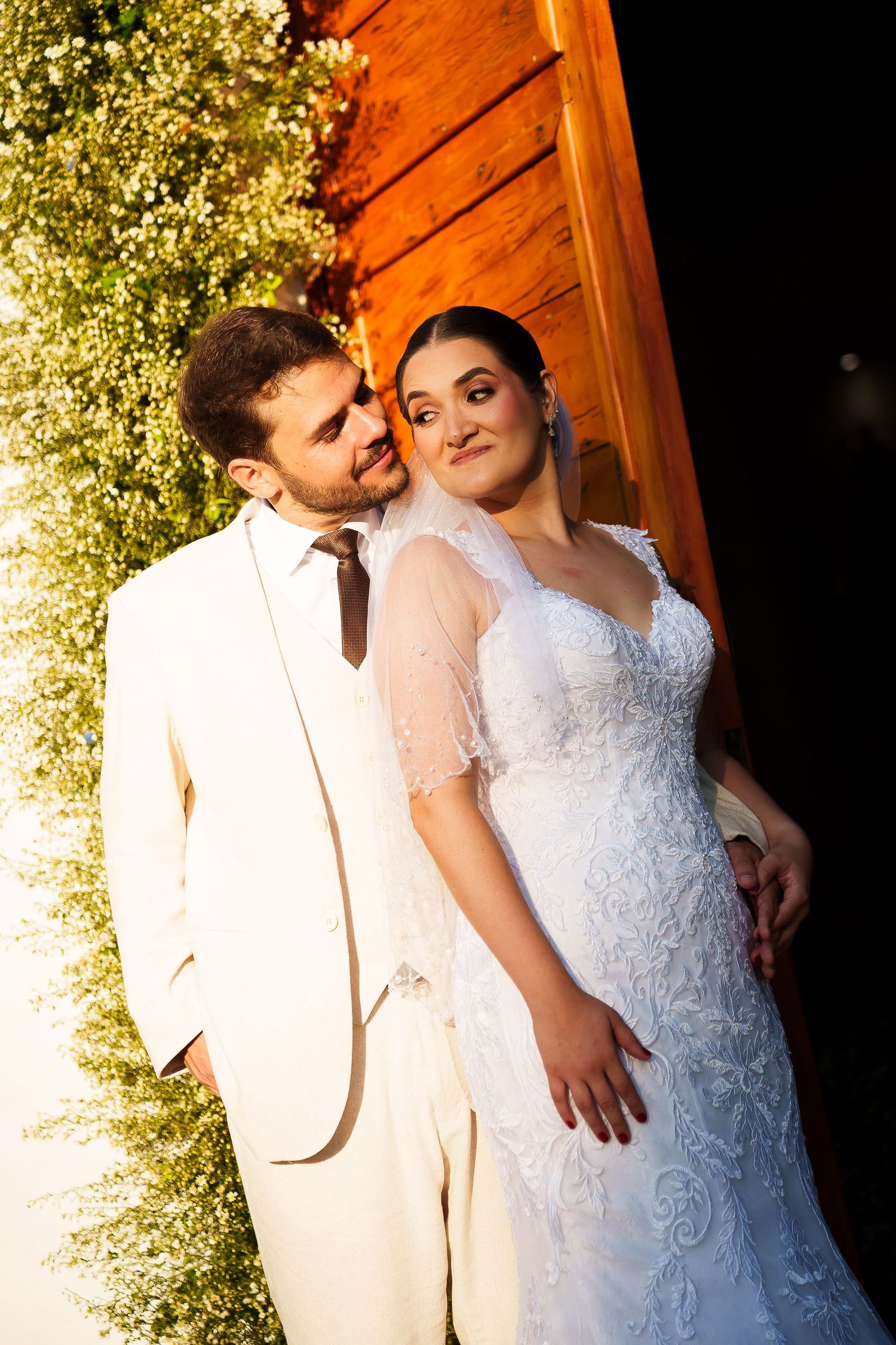 Bride in white lace gown and groom in ivory suit pose romantically near greenery and wooden door at night.