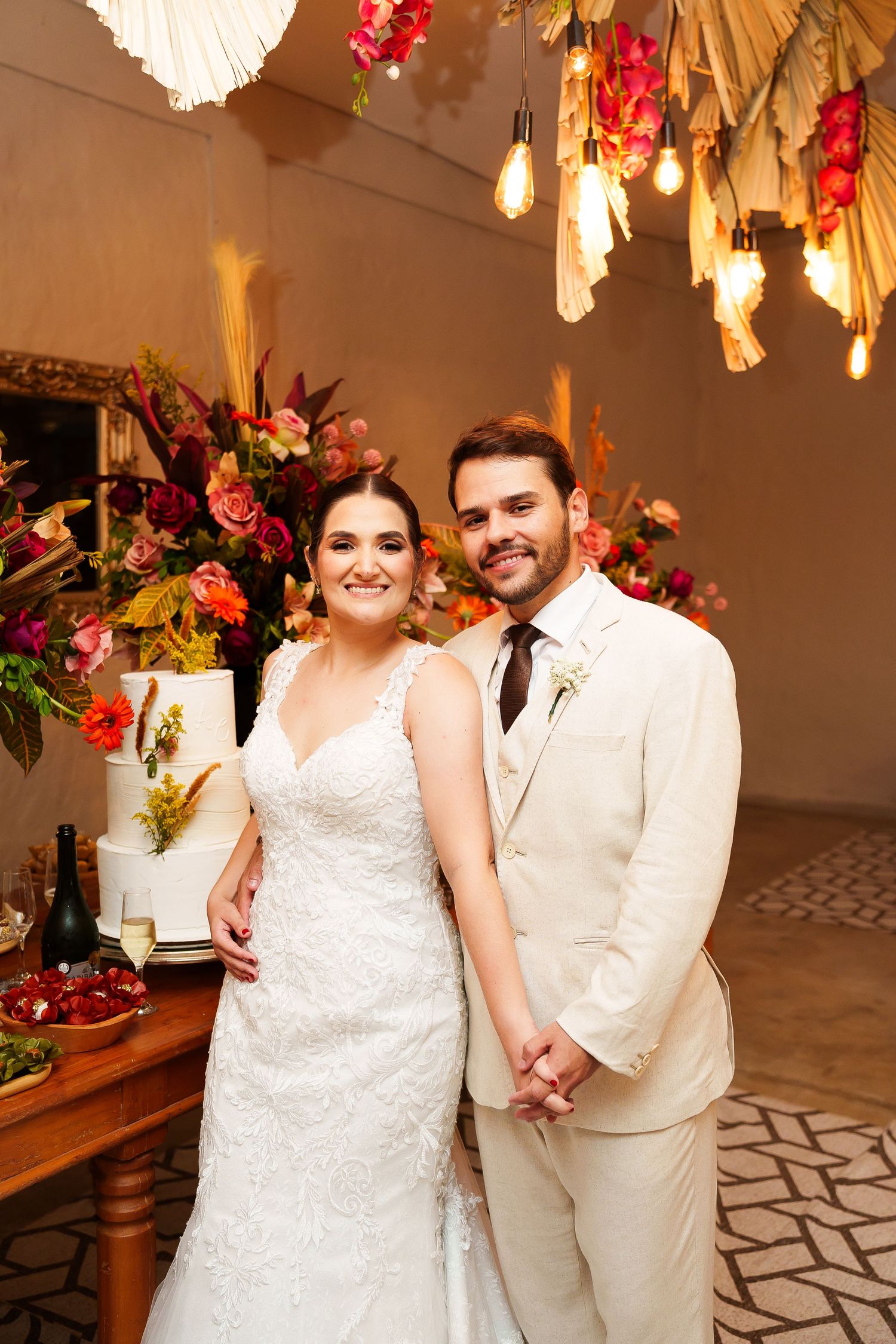 Bride in white lace gown and groom in cream suit pose together at elegant wedding reception with colorful floral backdrop.