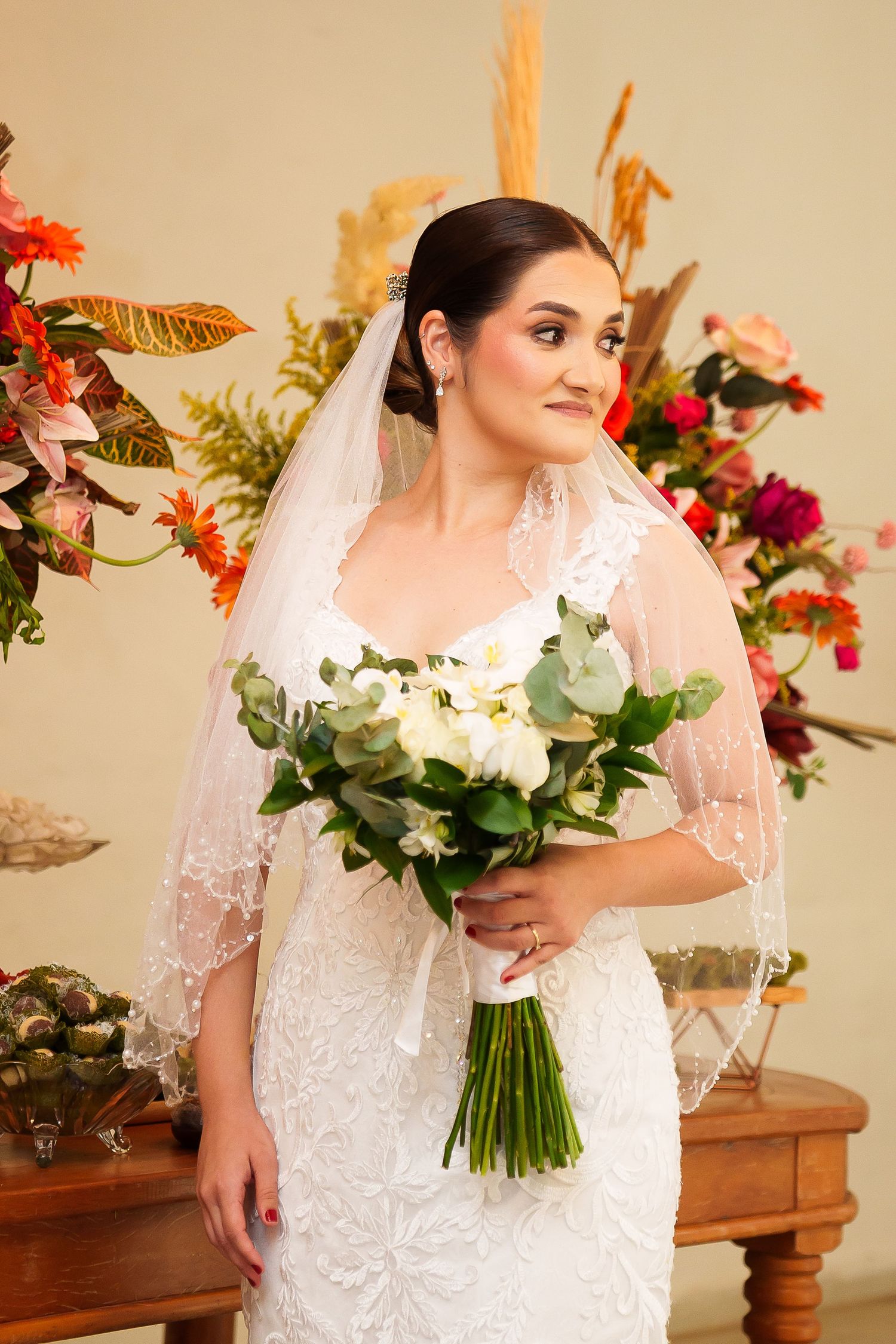Elegant bride in lace wedding dress holding white floral bouquet, wearing veil, posing near colorful flower arrangements.