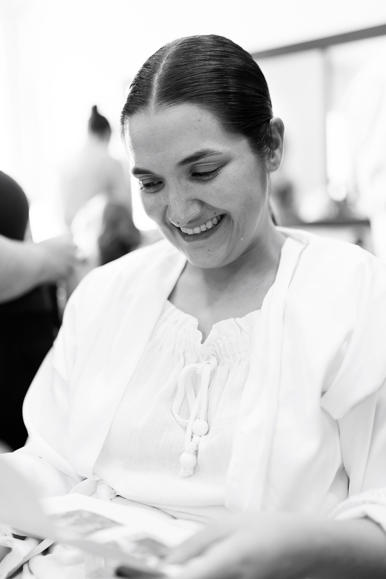 Black and white photo of a smiling woman in a white blouse looking down at papers in a bright room.
