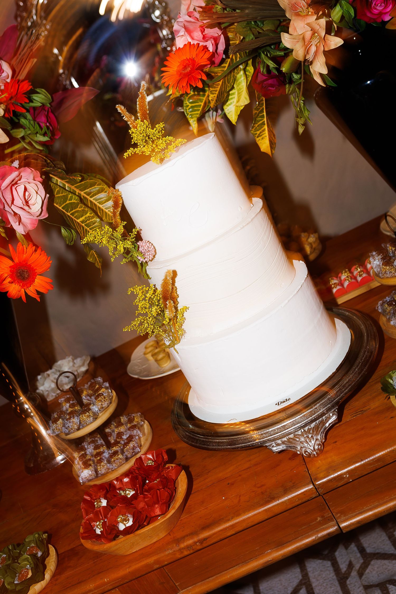 Elegant two-tier white wedding cake on a gold stand, surrounded by colorful flowers and desserts on a wooden table.