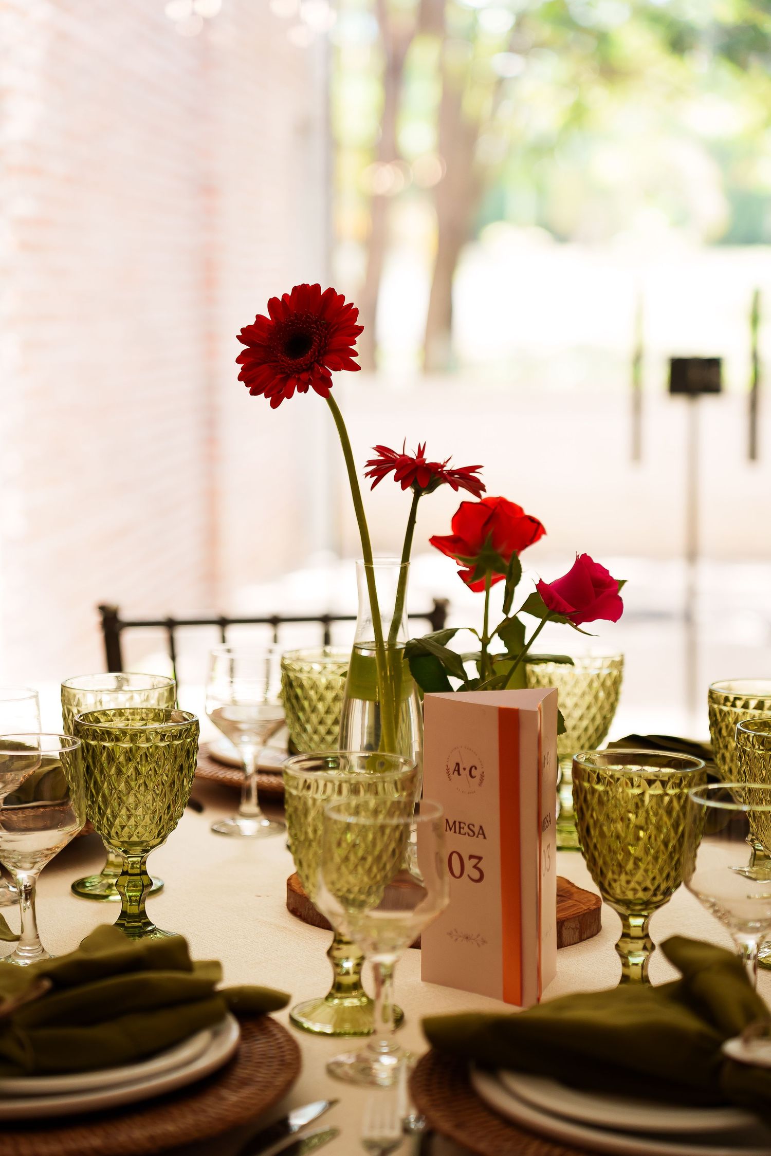 Elegant wedding table setting with red gerbera daisies, green goblets, white plates, and a Mesa 03 table number card.