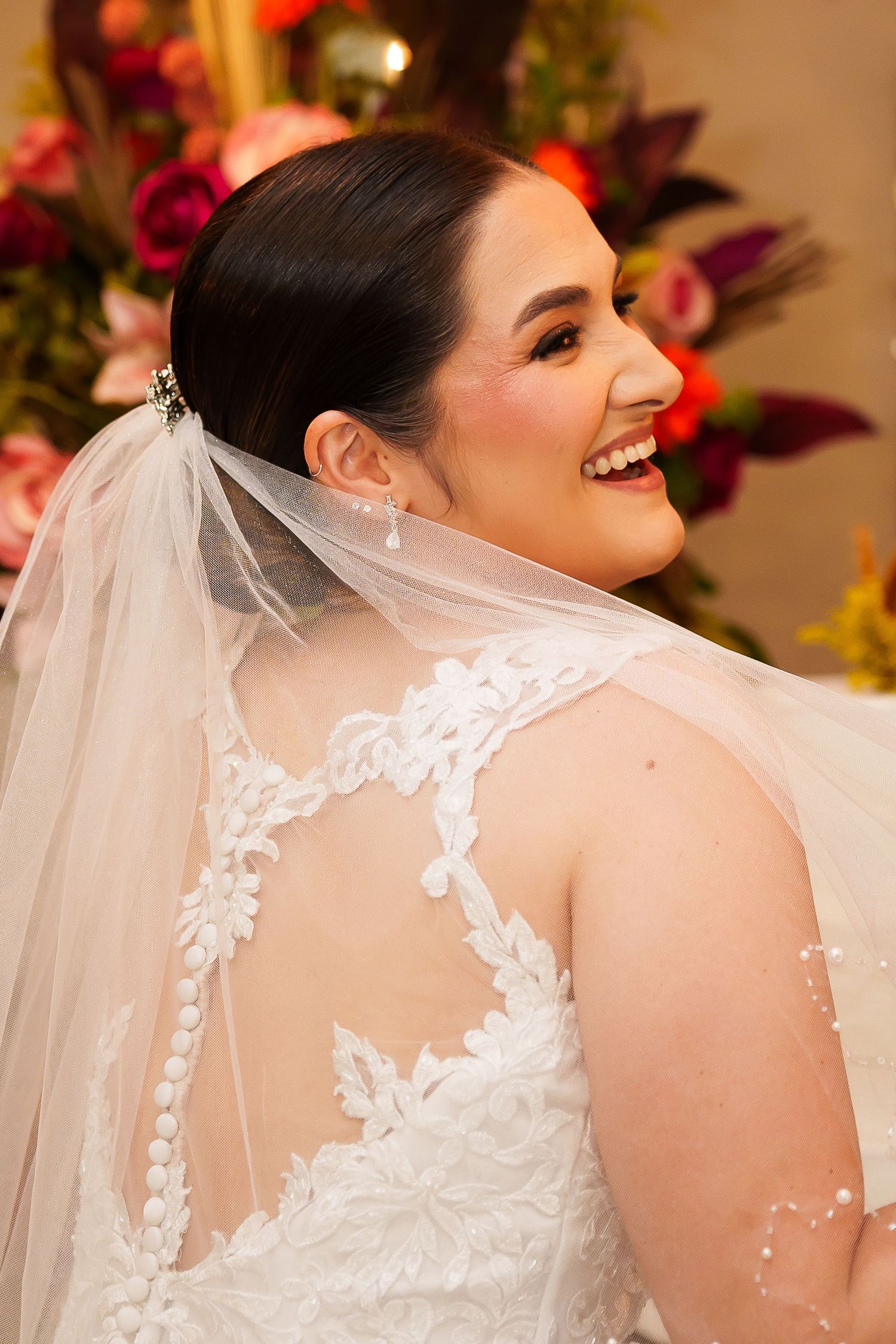 Smiling bride in white lace wedding dress with veil, showing intricate back detail, with floral arrangement in background.