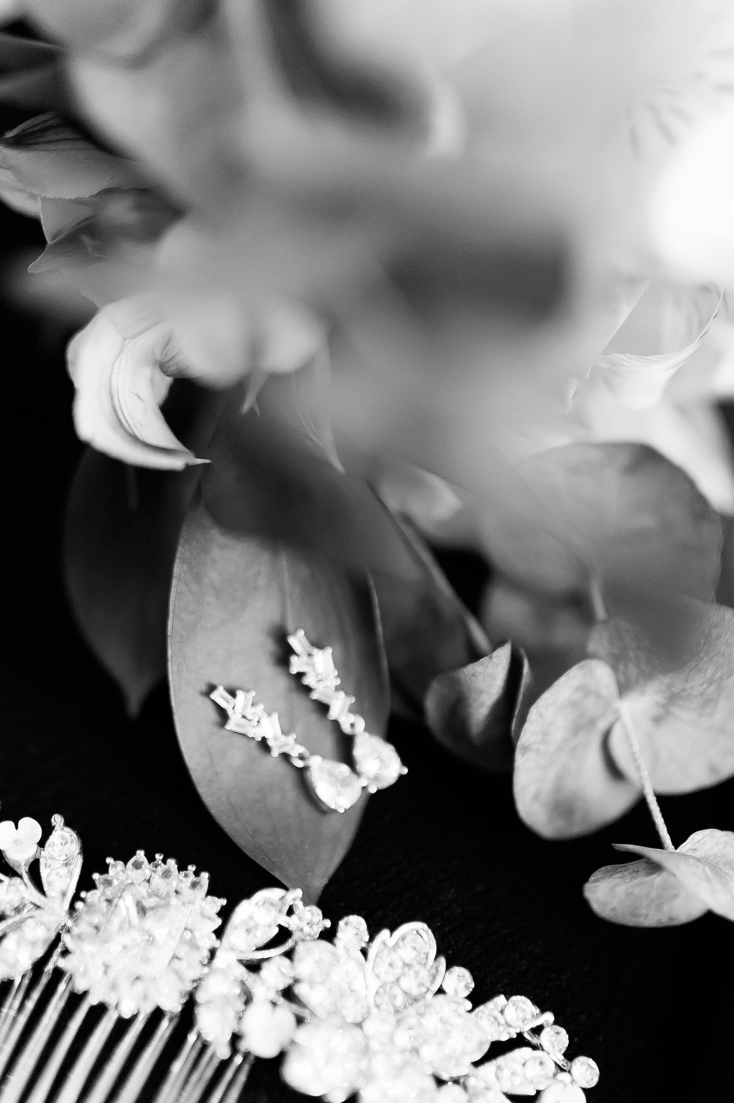 Black and white bridal accessories with floral hair comb, delicate earrings, and eucalyptus leaves on dark background.