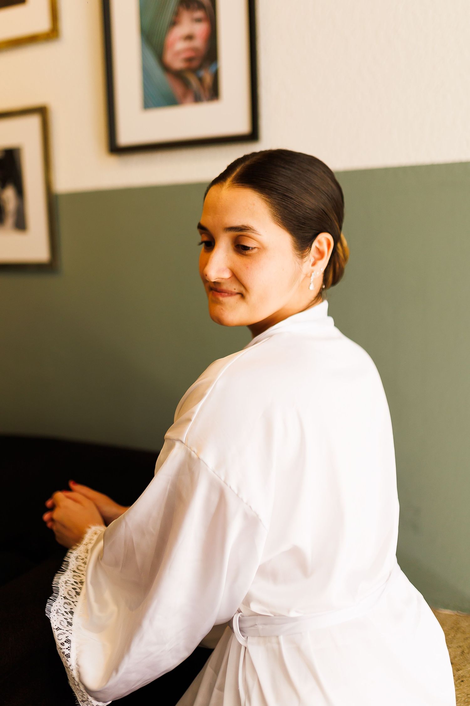 A smiling bride in a white lace robe sits gracefully, with framed artwork on a green wall behind her.