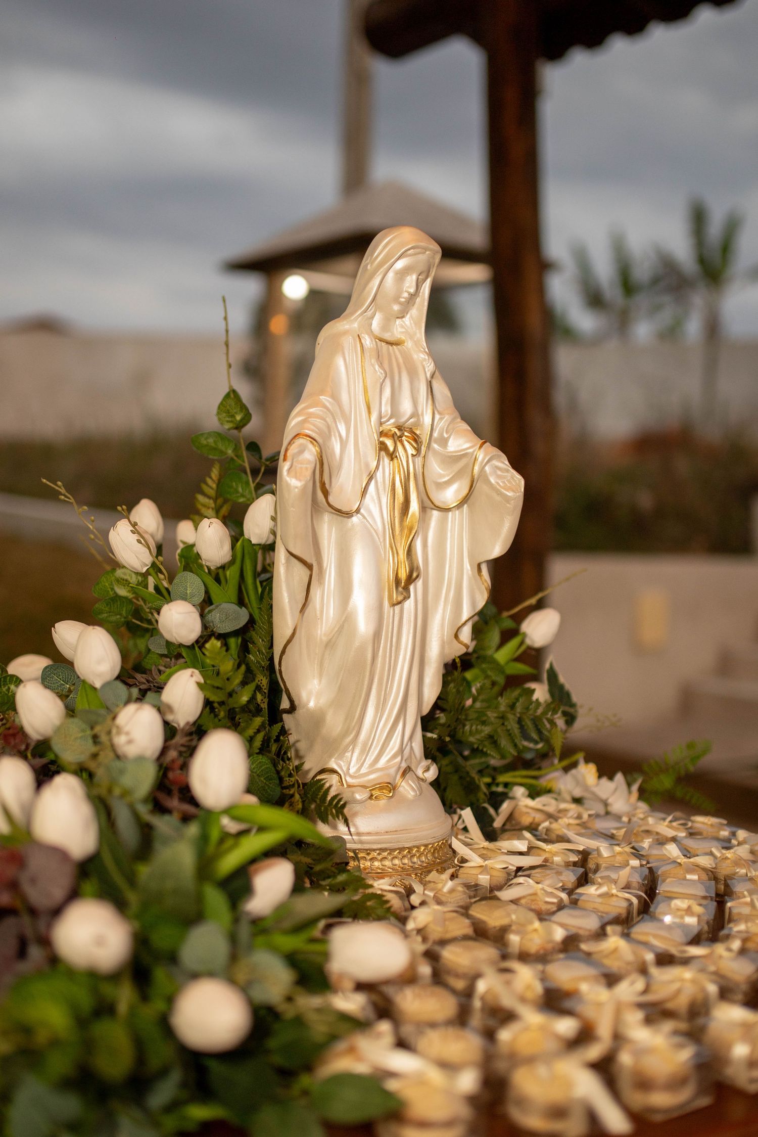 Pearl-finish Virgin Mary statue with gold accents surrounded by white roses and candles at an outdoor religious ceremony.