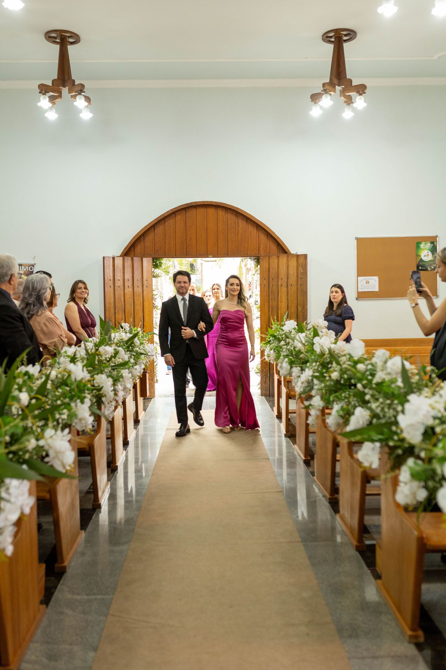 A couple walks down a church aisle, woman in magenta gown, man in suit, flanked by white floral arrangements on wooden pews.