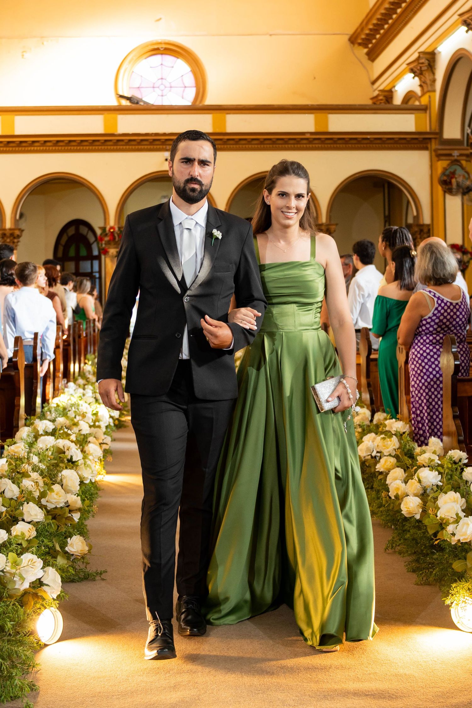 Elegant couple walking down a flower-lined church aisle, man in black tuxedo, woman in green satin gown.