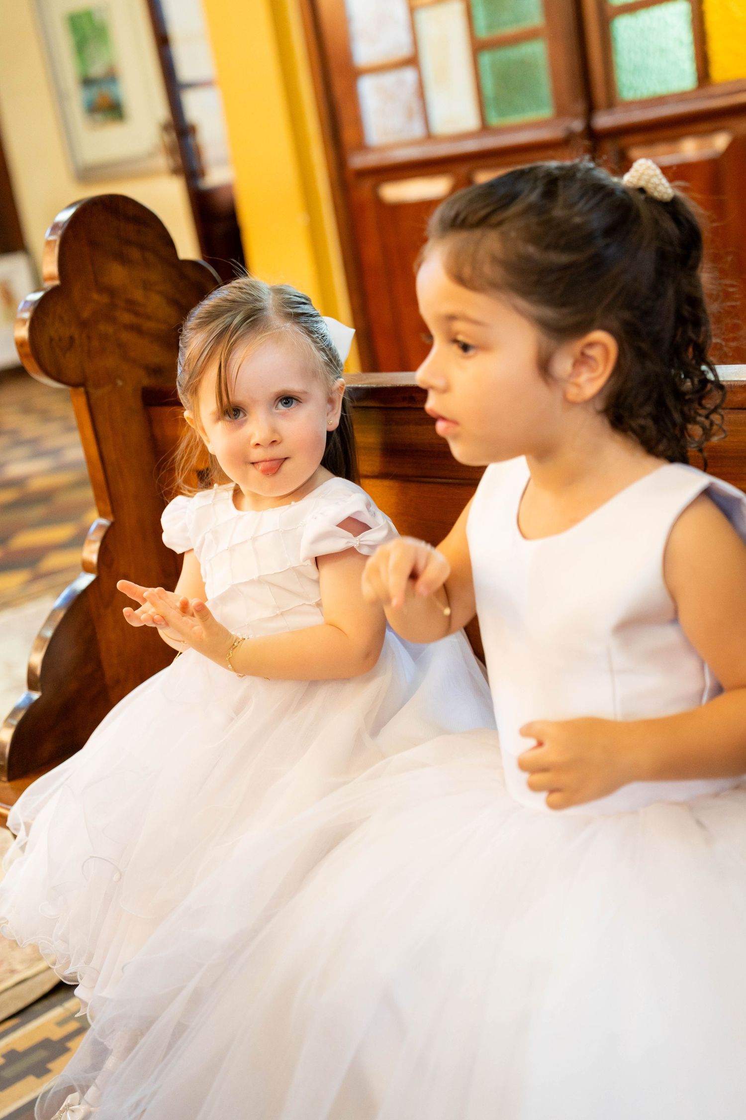 Two young girls in white dresses sitting on a wooden church pew, likely flower girls at a wedding ceremony.